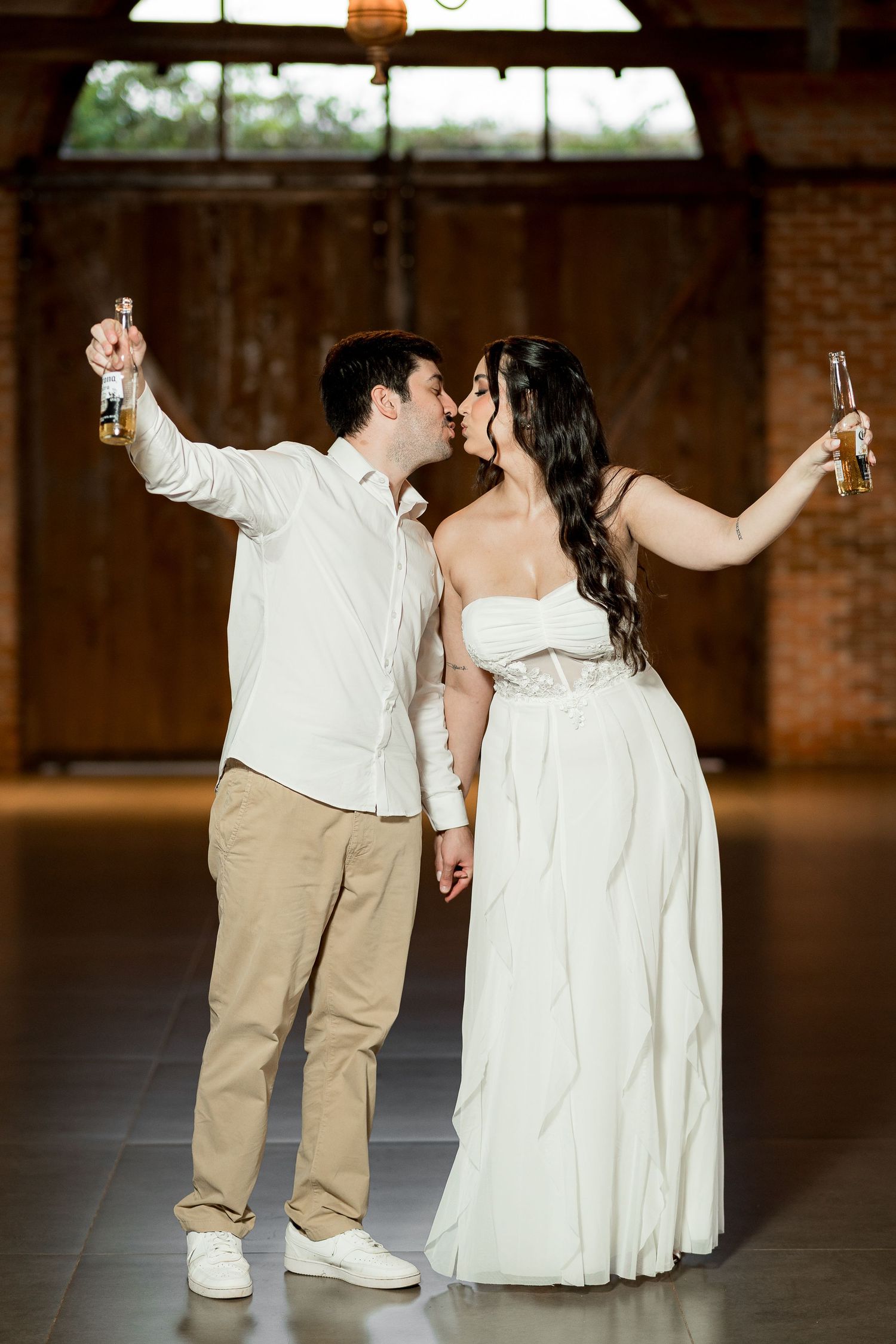 Bride and groom kissing while raising beer bottles in a rustic brick venue during their wedding celebration.