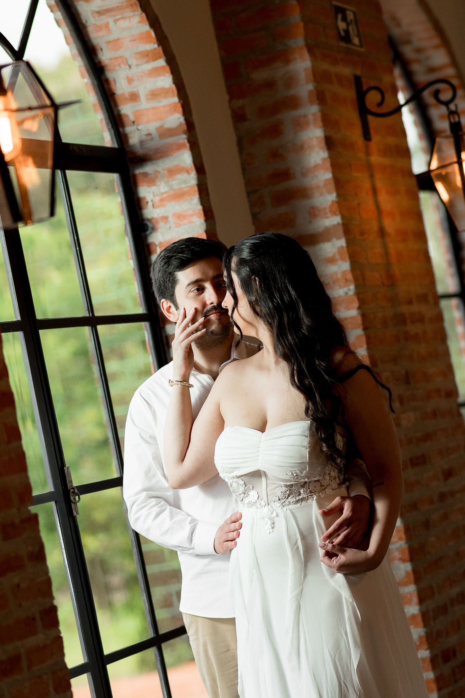 A bride and groom share a romantic moment by arched windows in a rustic brick venue, the bride in a white strapless gown.