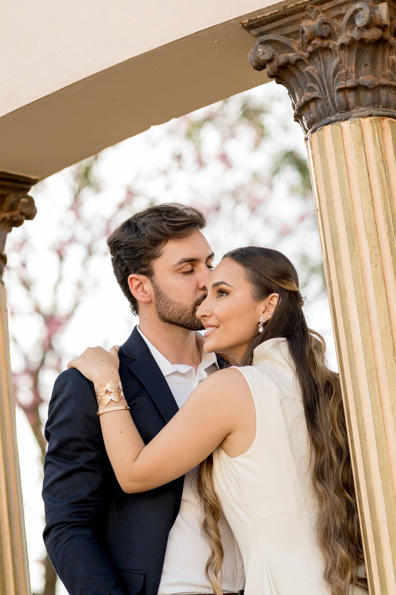 Couple sharing a romantic kiss between ornate columns, woman in white dress smiling, man in navy suit outdoors.