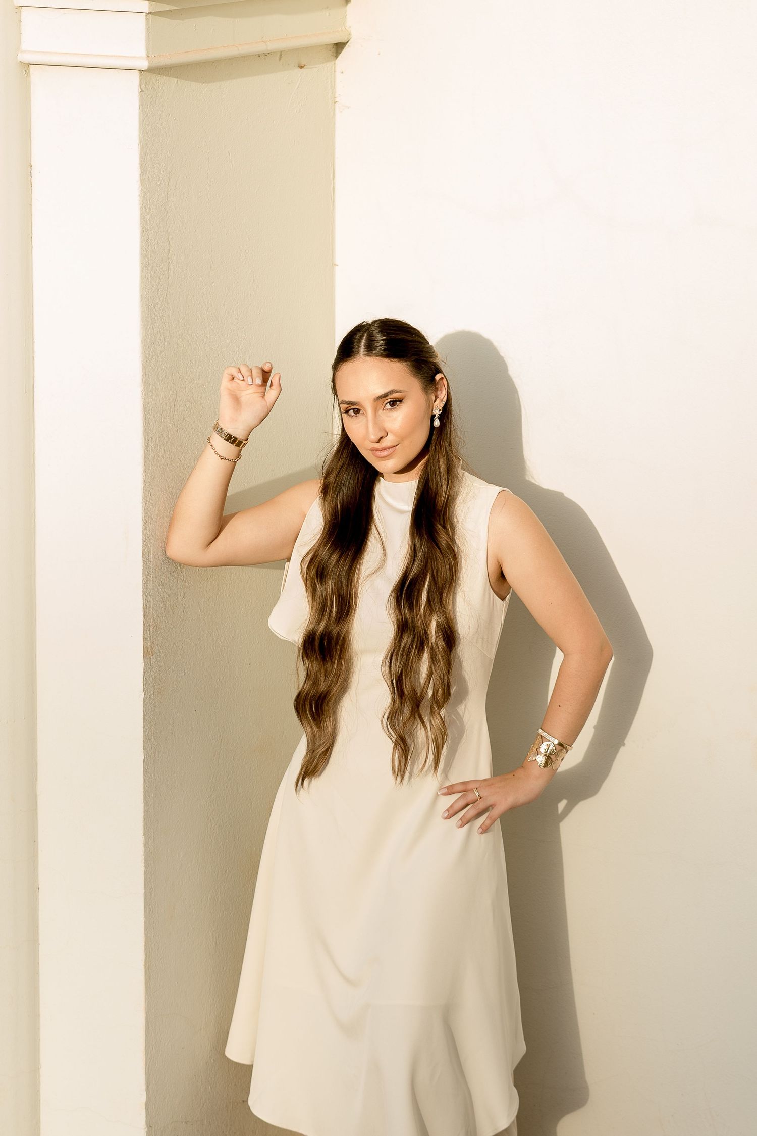 Woman in white sleeveless dress with long wavy hair poses gracefully against a white wall with column.
