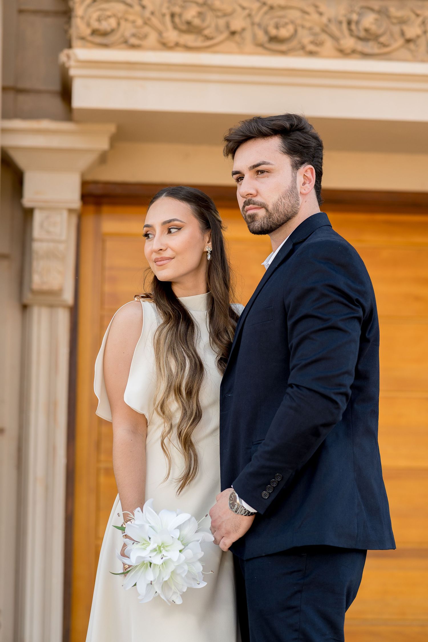 Elegant bride in white dress holding lily bouquet stands with groom in navy suit at ornate building.