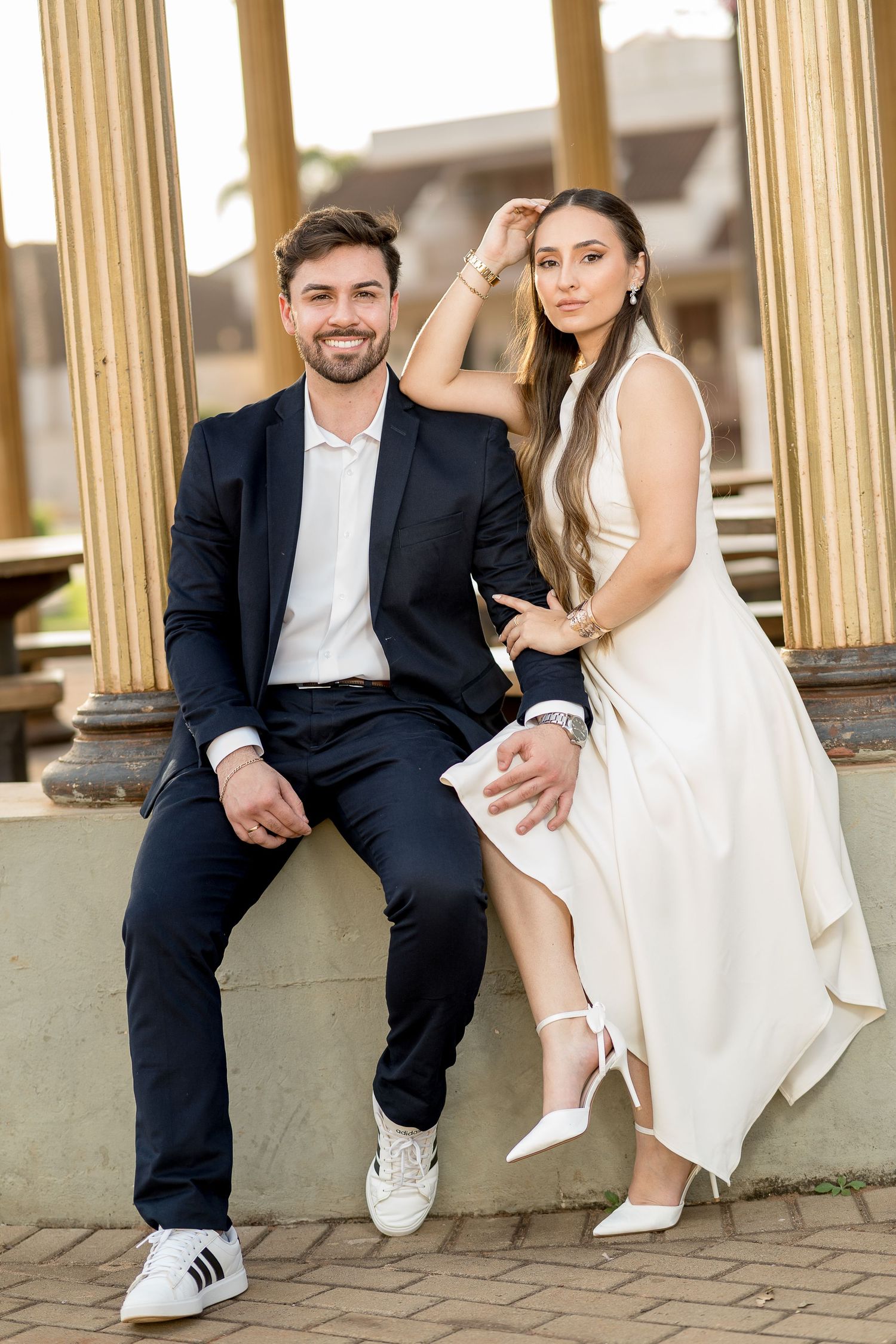 Elegant couple posing outdoors, man in black suit with white sneakers, woman in white dress with heels, near classical columns.