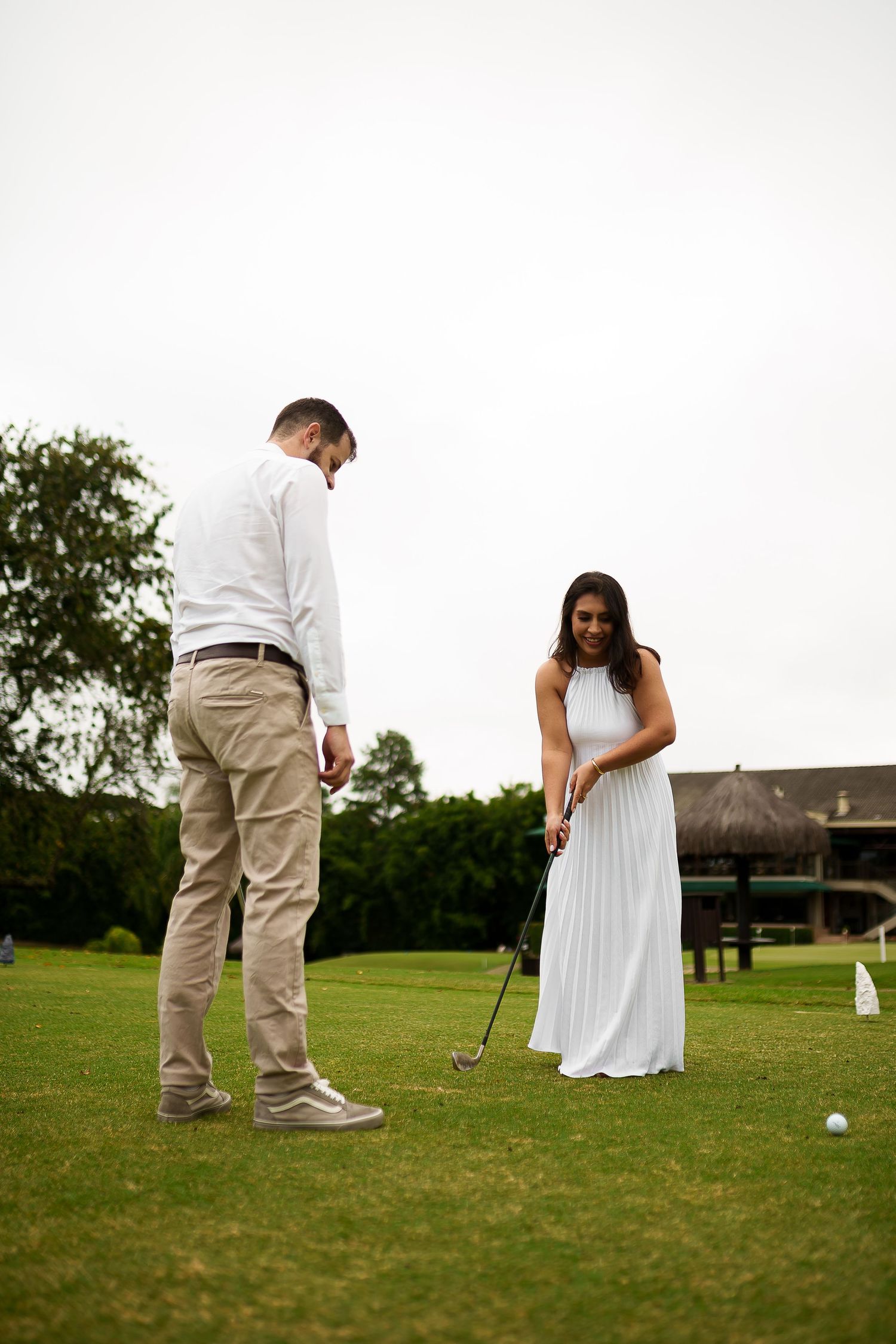 Couple playing golf on a green course, woman in white dress holding club, man in white shirt and khaki pants nearby.