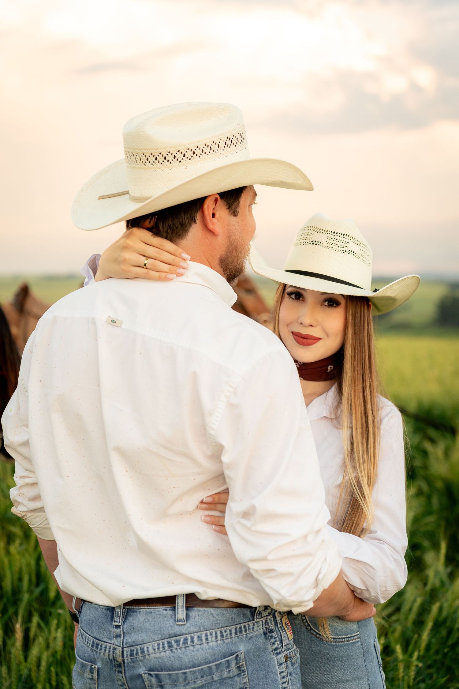 Couple in white shirts and cowboy hats embracing in a green field at sunset, romantic western style photo.
