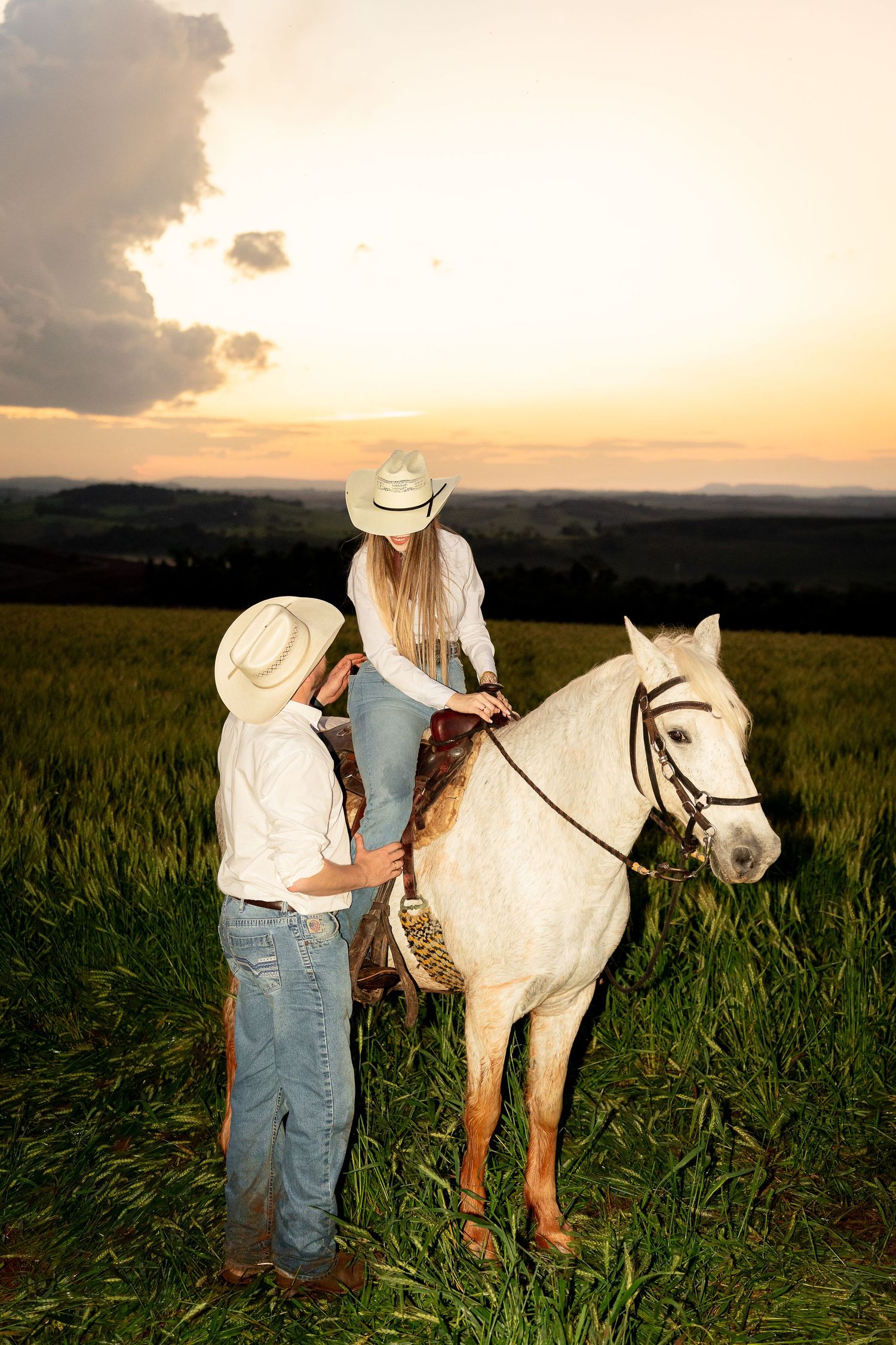 A woman in a cowboy hat rides a white horse in a green field at sunset, assisted by a man in a white shirt.