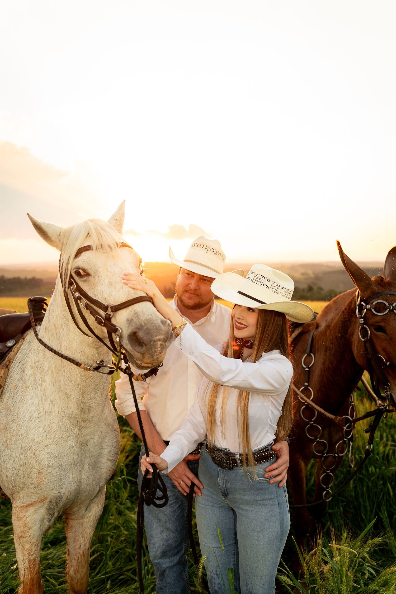 Couple in cowboy hats petting a white horse at sunset in a green field with a brown horse behind them.