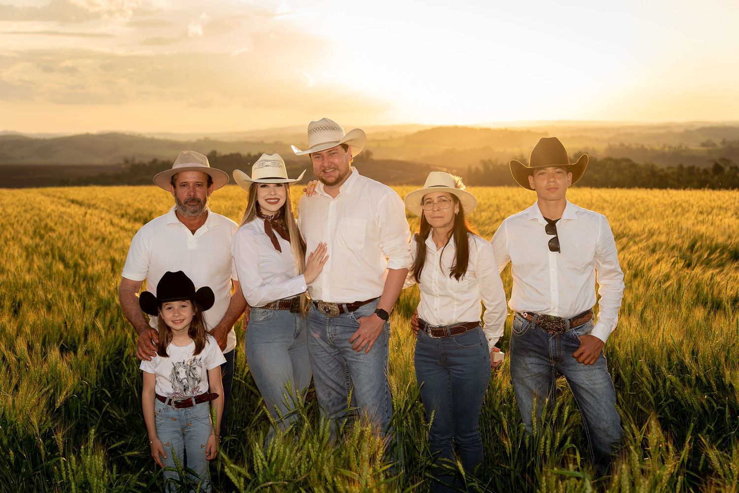 Six people in cowboy hats and jeans pose in a golden wheat field at sunset on a farm.