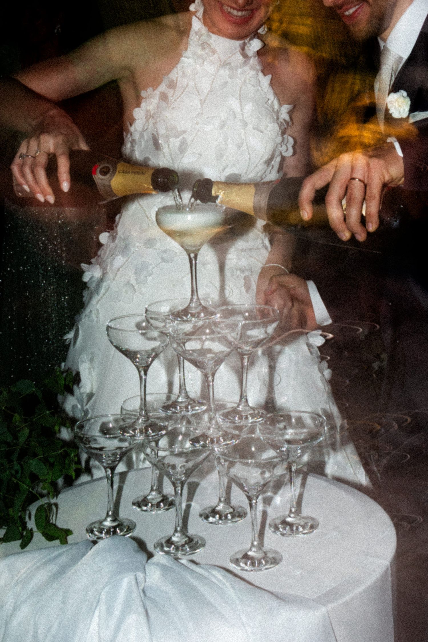 Bride and groom pour champagne over a tiered tower of coupe glasses at their wedding celebration.