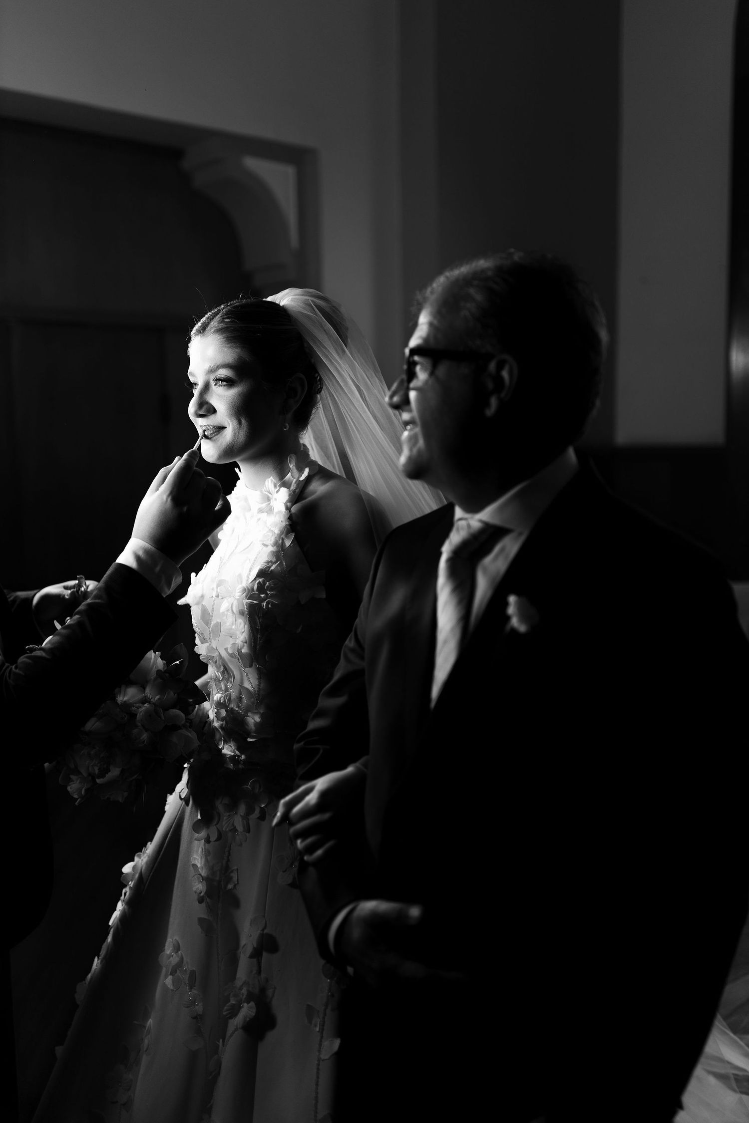 Black and white photo of a smiling bride in lace gown walking with her father on her wedding day.