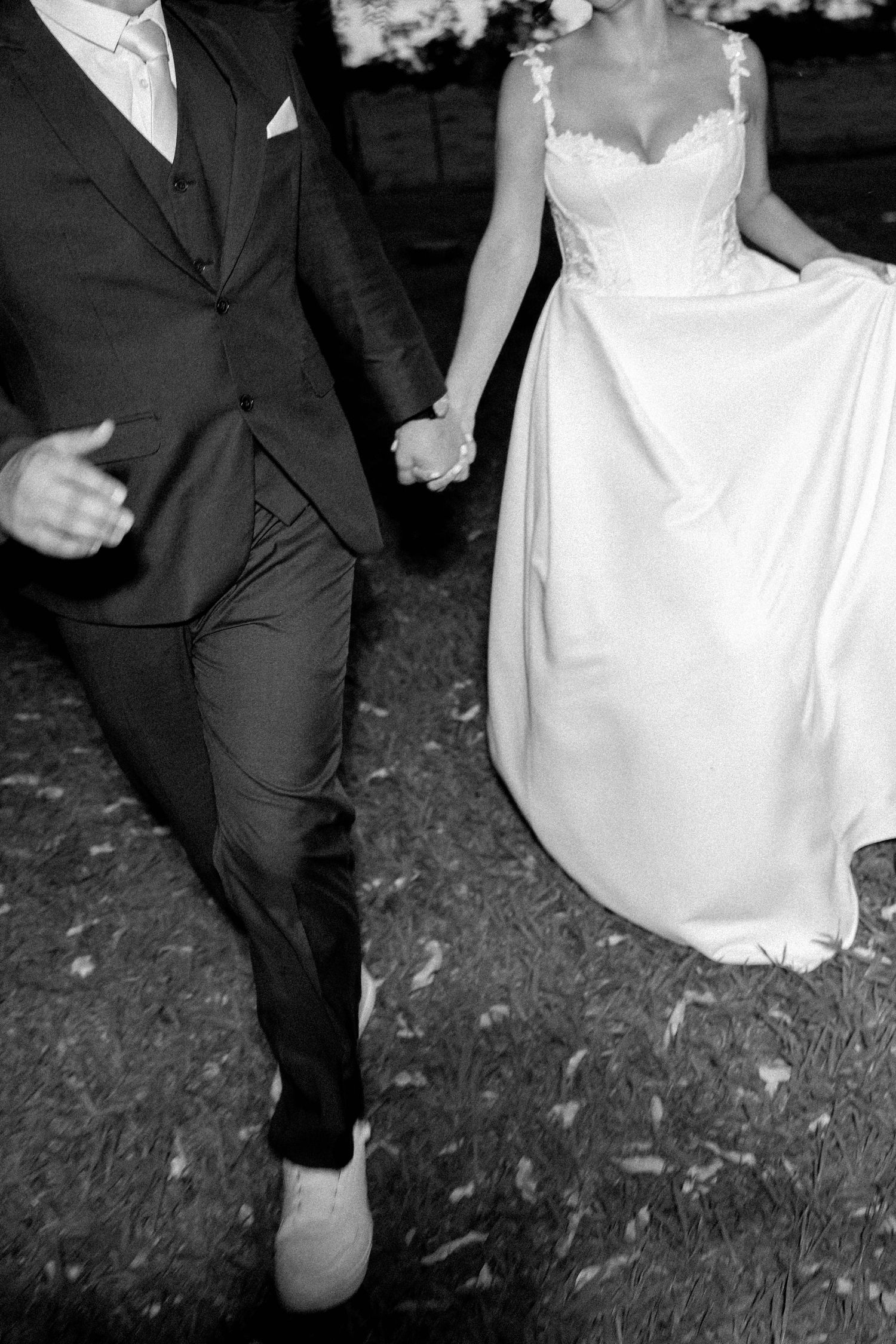Black and white photo of bride and groom holding hands, walking together outdoors in wedding attire.