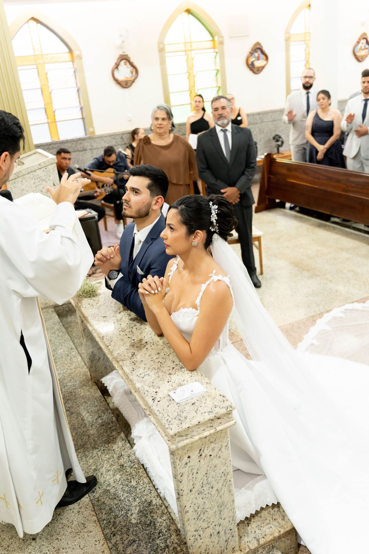 Bride and groom kneeling at church altar during Catholic wedding ceremony, guests standing in pews behind them.