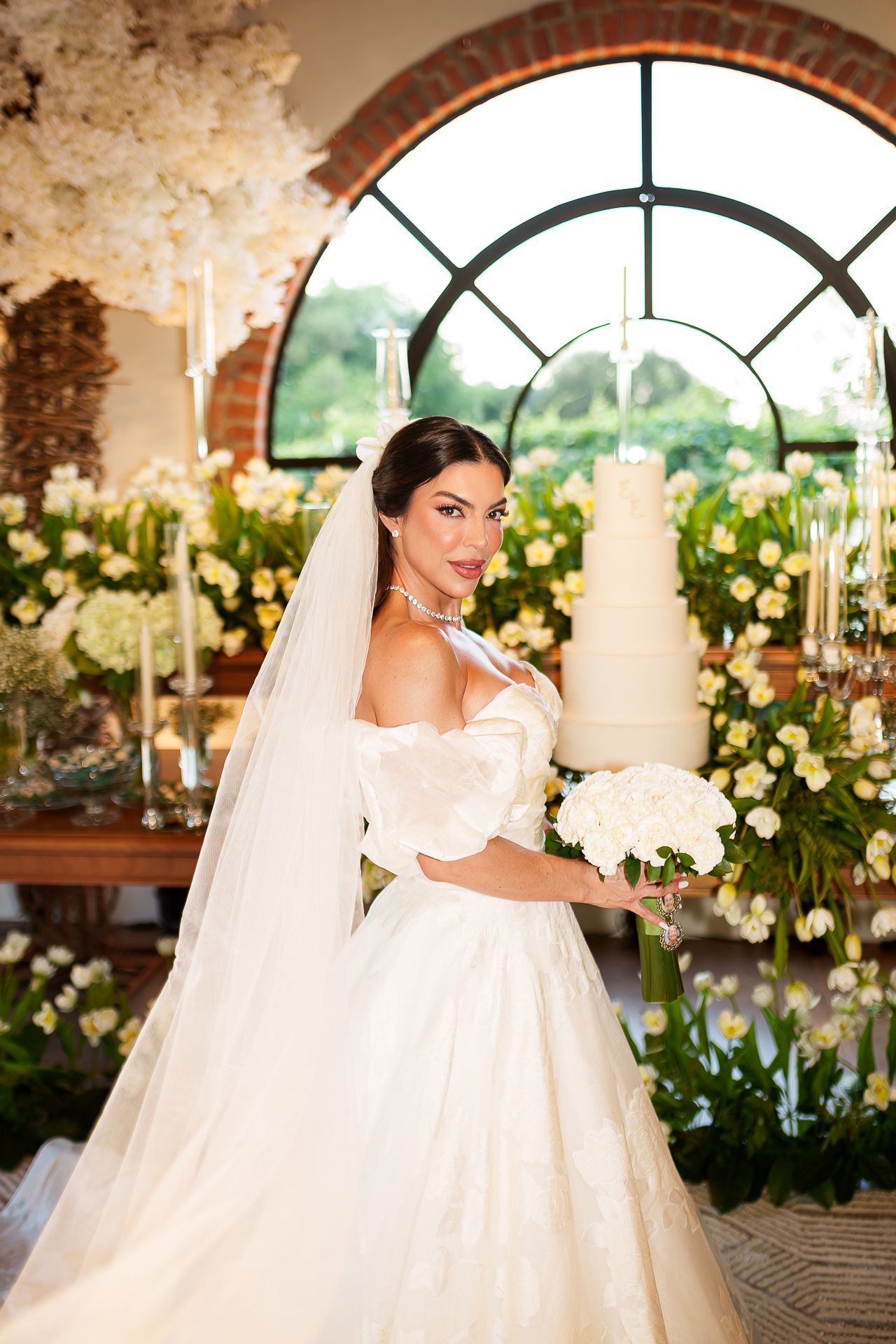 Elegant bride in off-shoulder white gown and veil holds bouquet before tiered wedding cake and floral arch.
