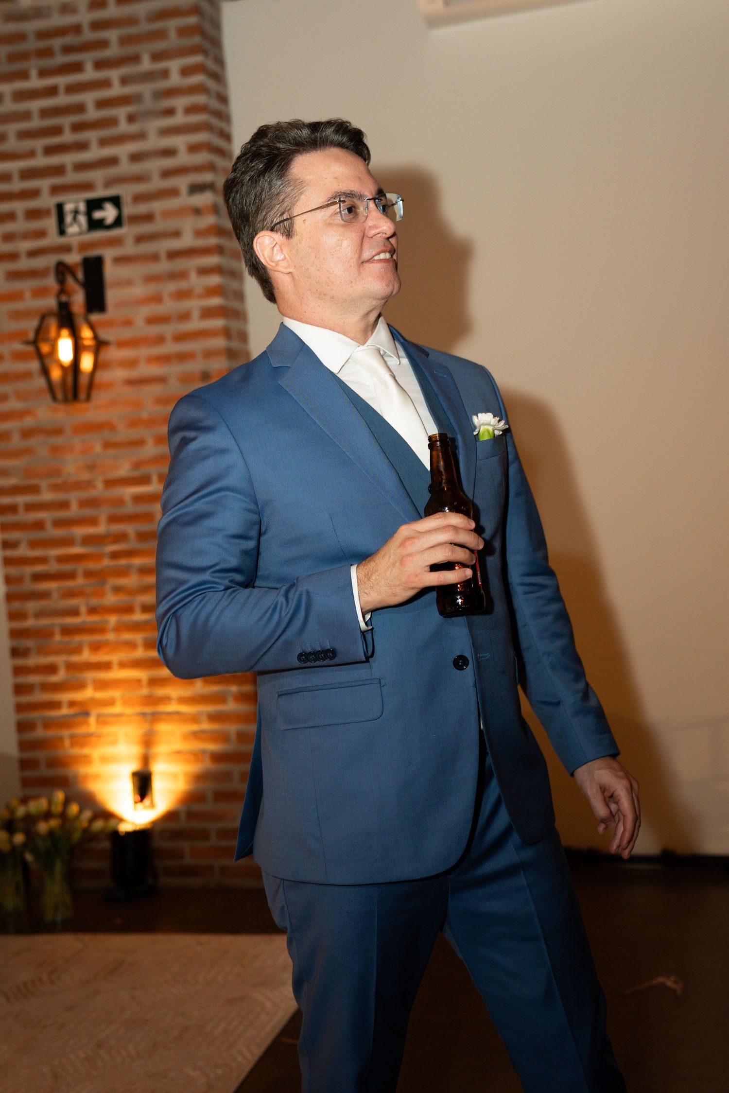 Man in blue suit holding beer bottle at elegant wedding reception with brick wall background.