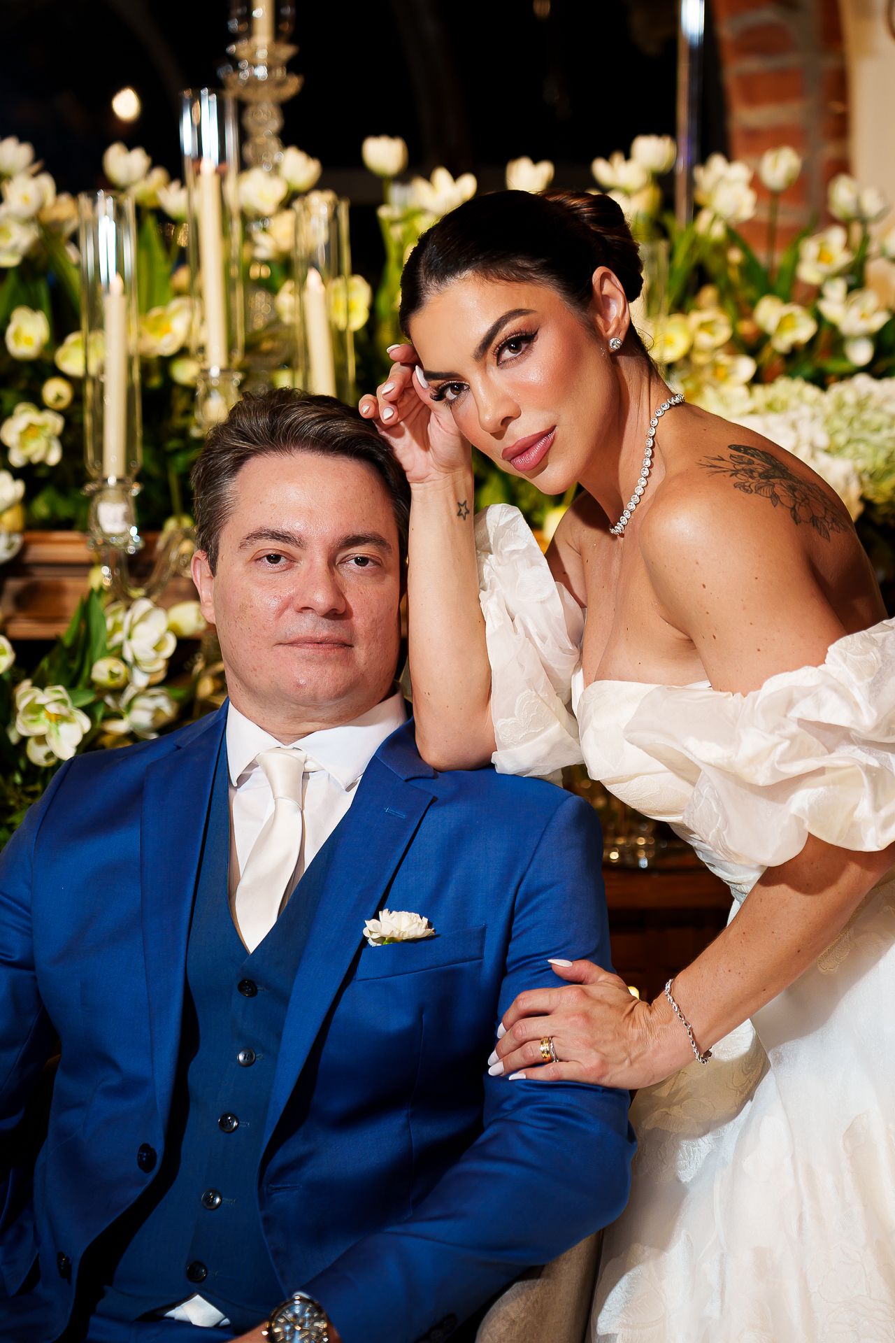 Elegant wedding couple posing together, groom in blue suit, bride in white off-shoulder gown with pearl necklace.