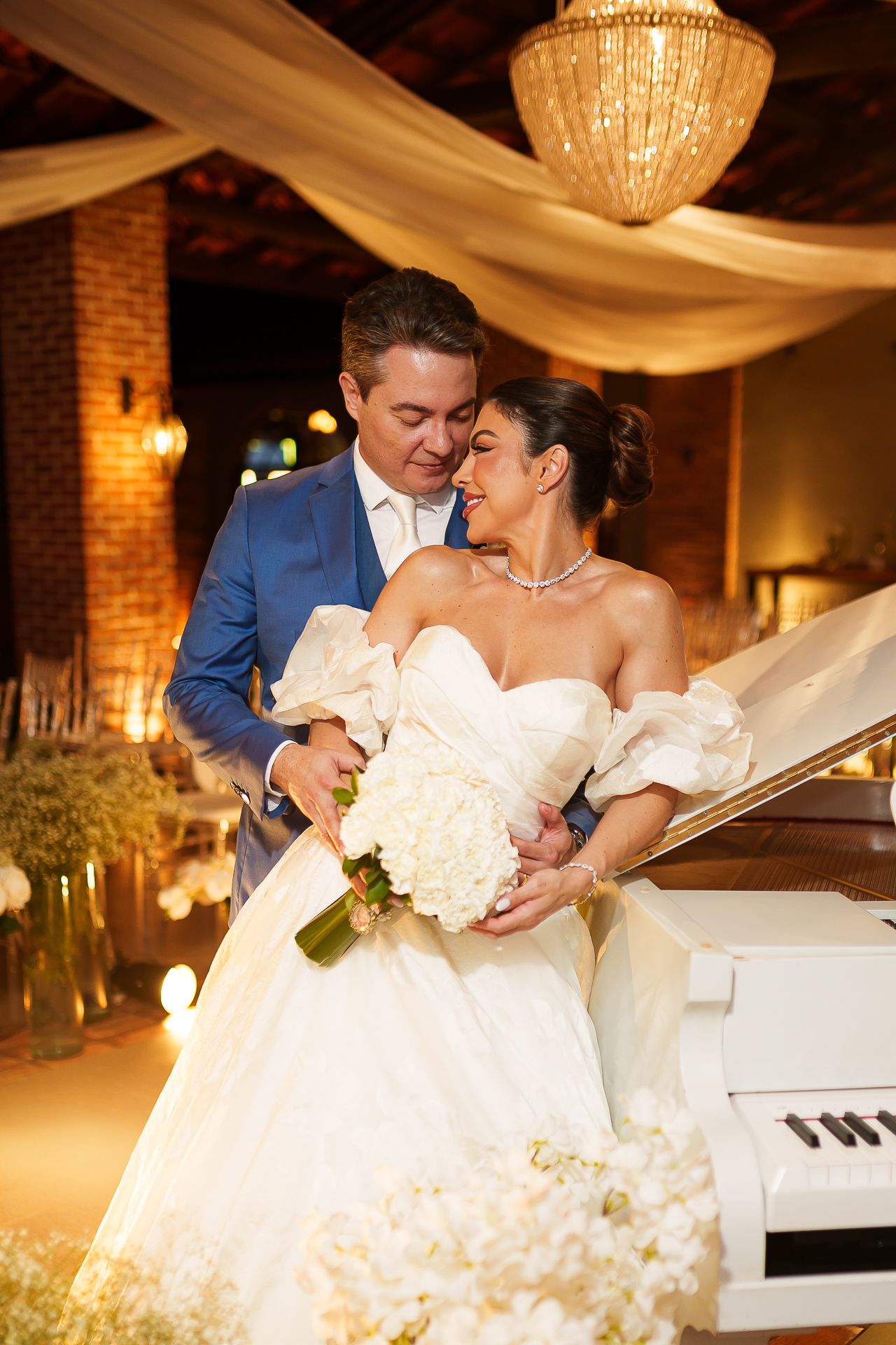 Groom in blue suit embraces bride in white ball gown holding bouquet by a white grand piano.