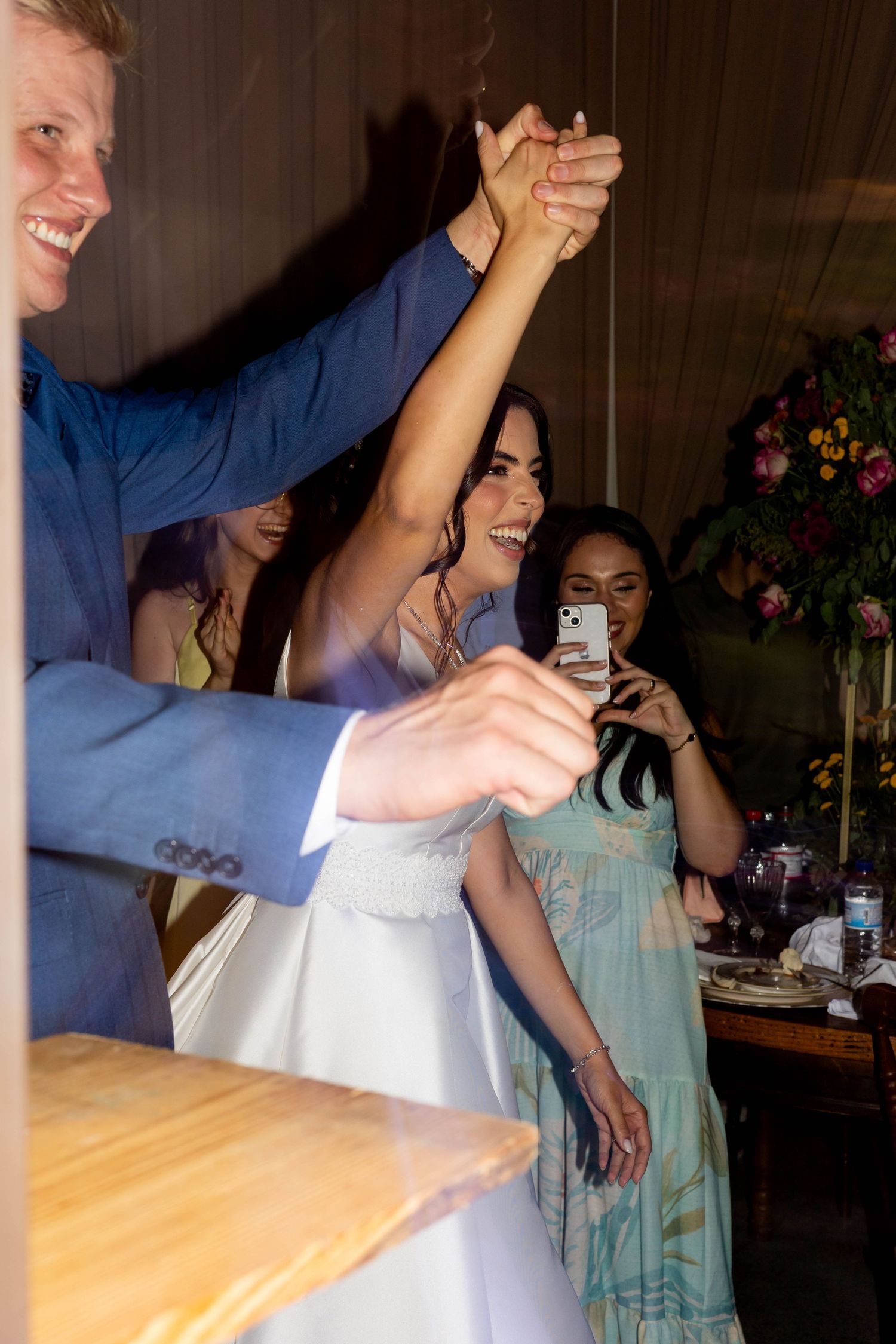 Bride and groom dancing joyfully at wedding reception while guest captures moment on smartphone.