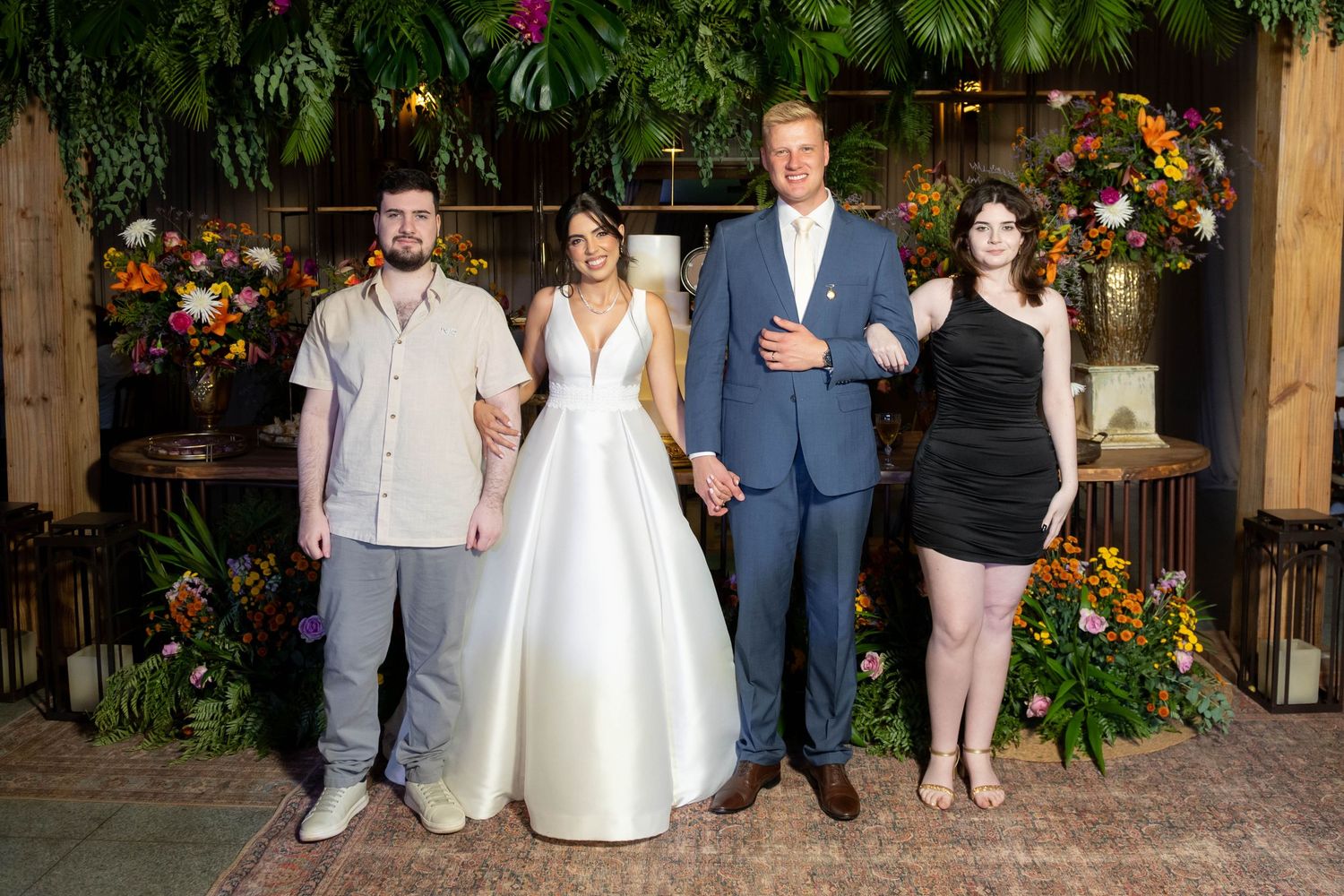 Bride and groom pose with two guests amid lush floral backdrop at elegant indoor wedding reception.