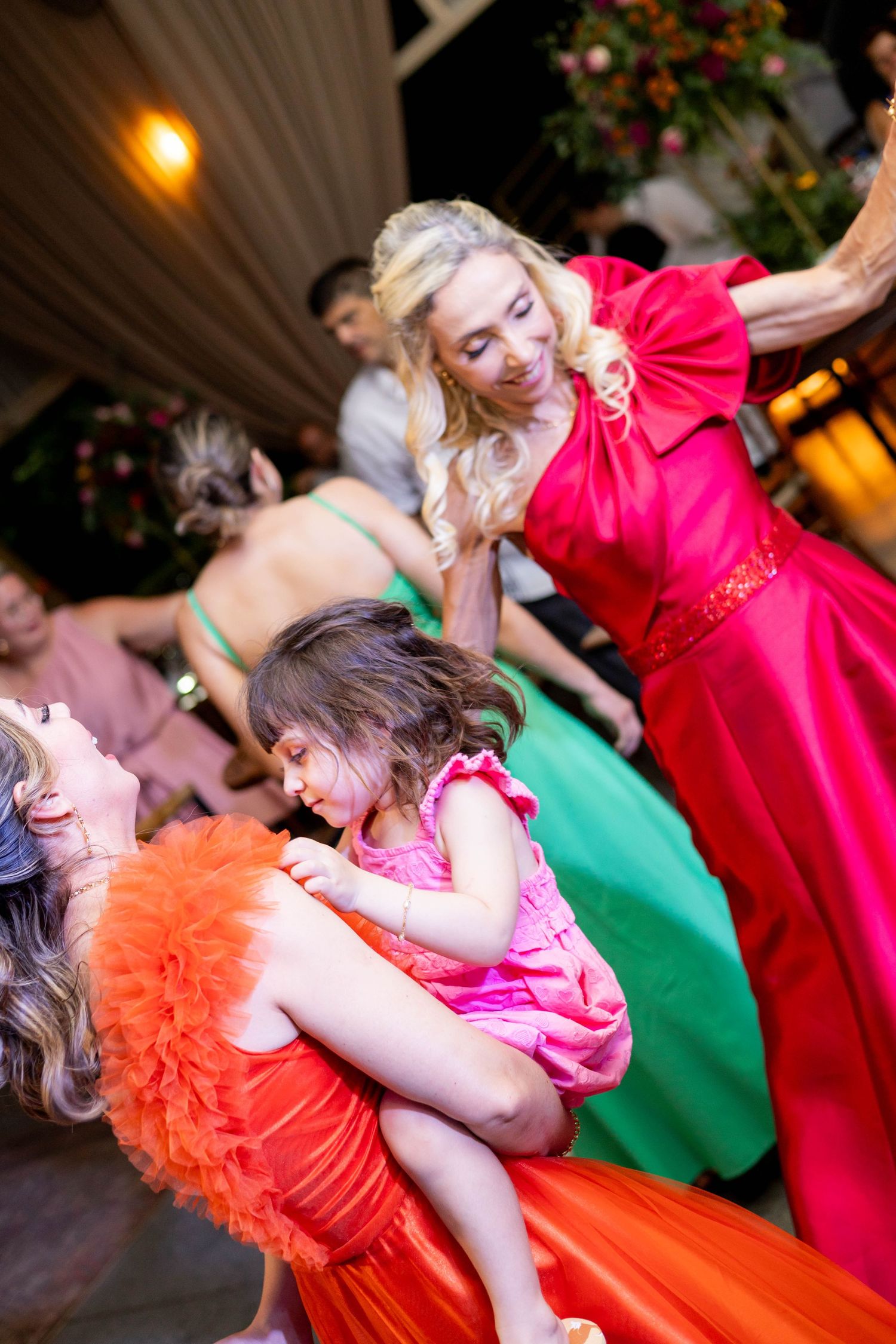 Women in colorful gowns dance joyfully at a festive event, with a blonde woman in red lifting a child.