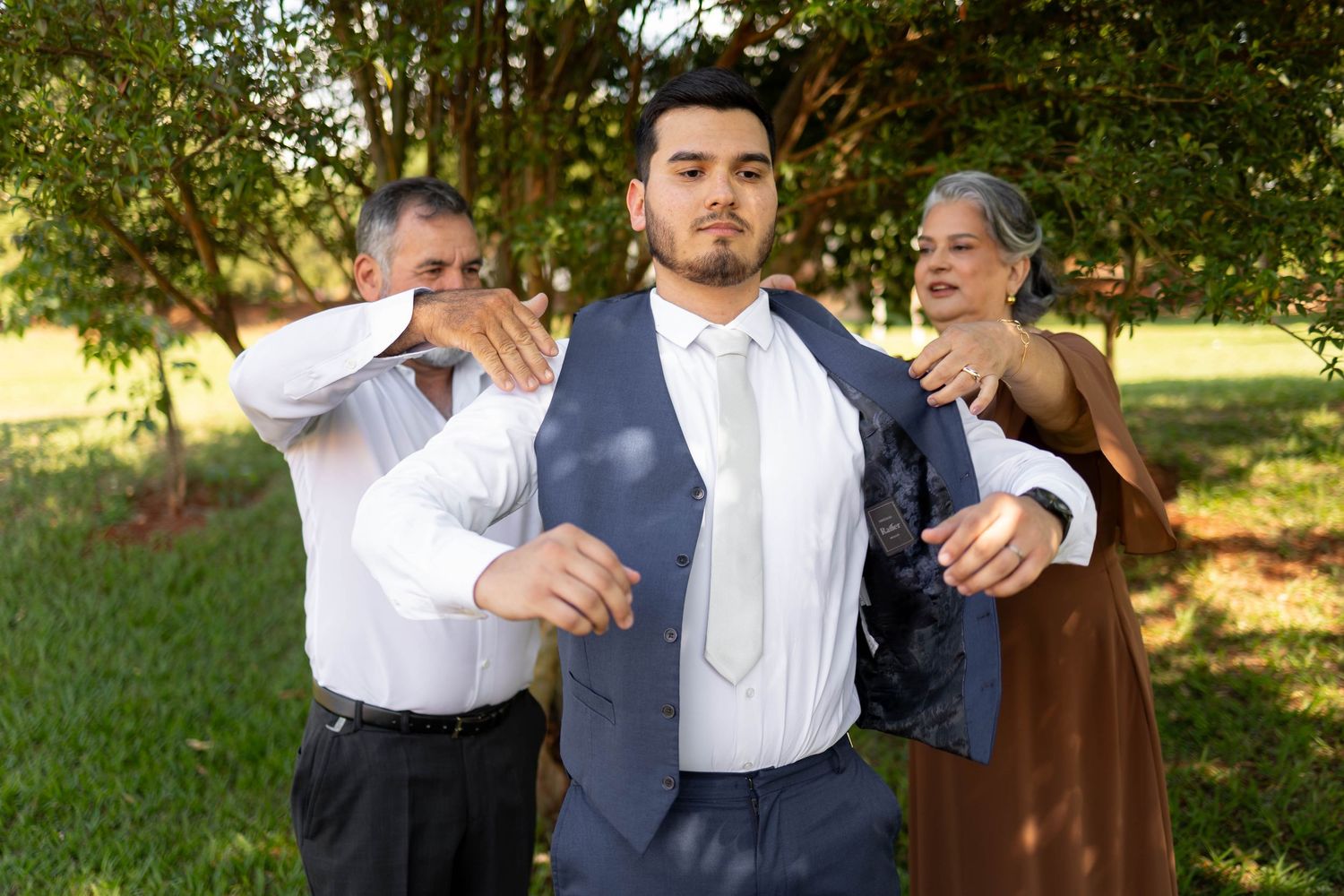 Young groom in blue vest and tie being helped by parents outdoors before wedding ceremony.