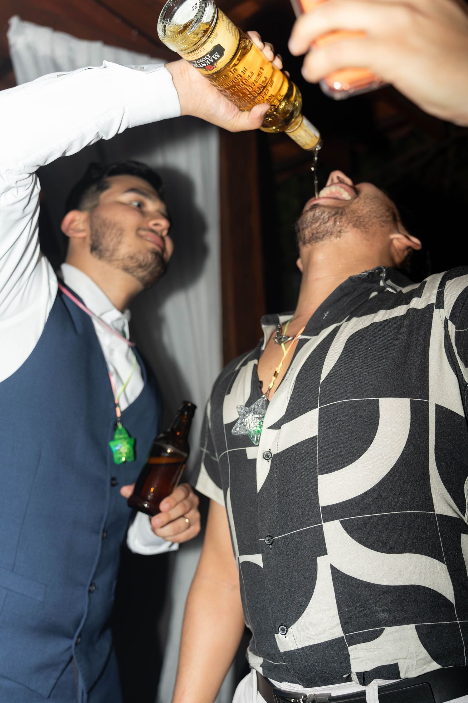 Two men at a party, one pouring liquor from a bottle into the other's mouth while holding a beer.