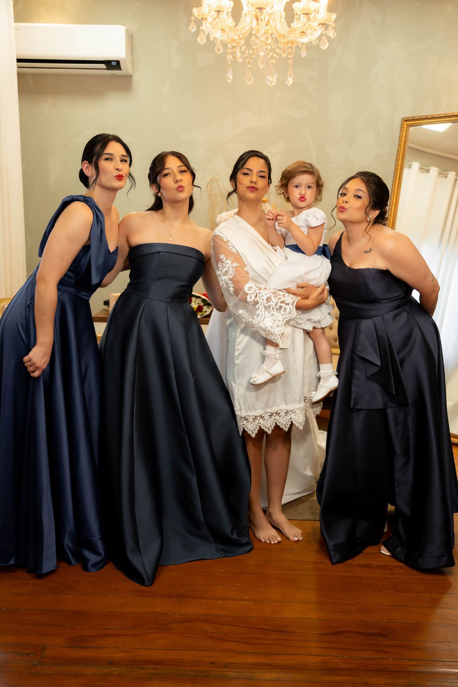 Bride in white robe holds toddler surrounded by three bridesmaids in navy satin gowns, posing playfully indoors.