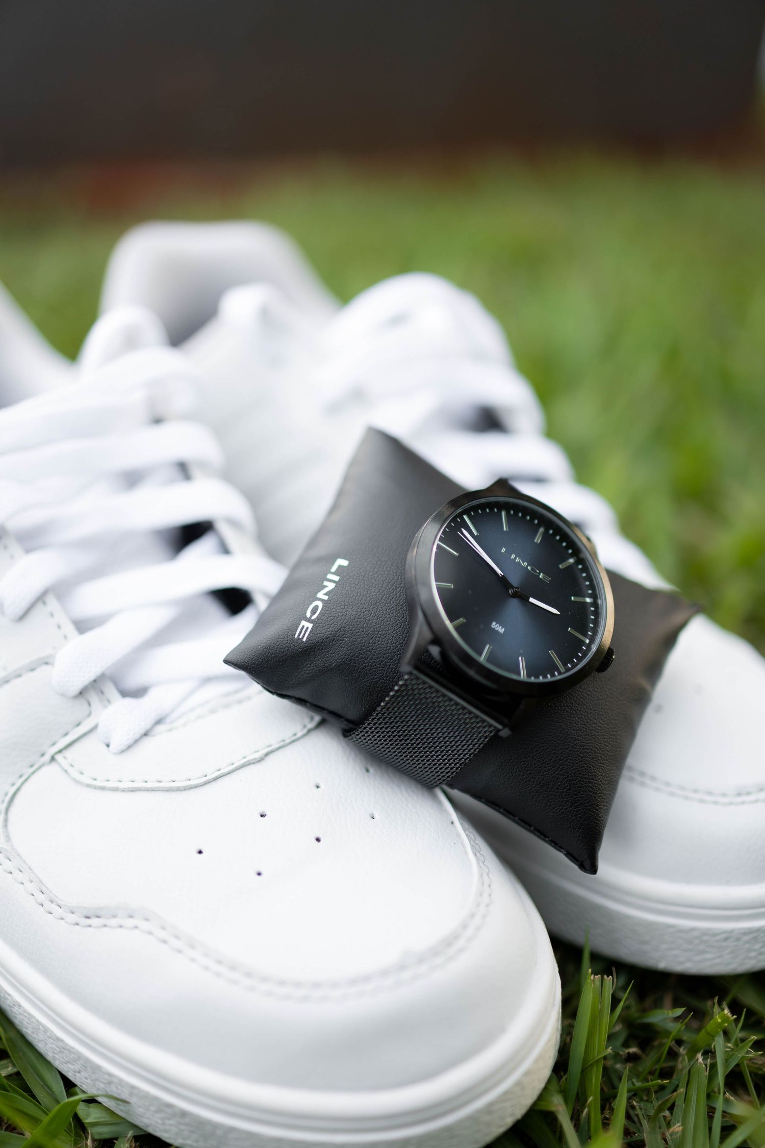 Black Lince watch with mesh band resting on white sneakers placed on green grass.