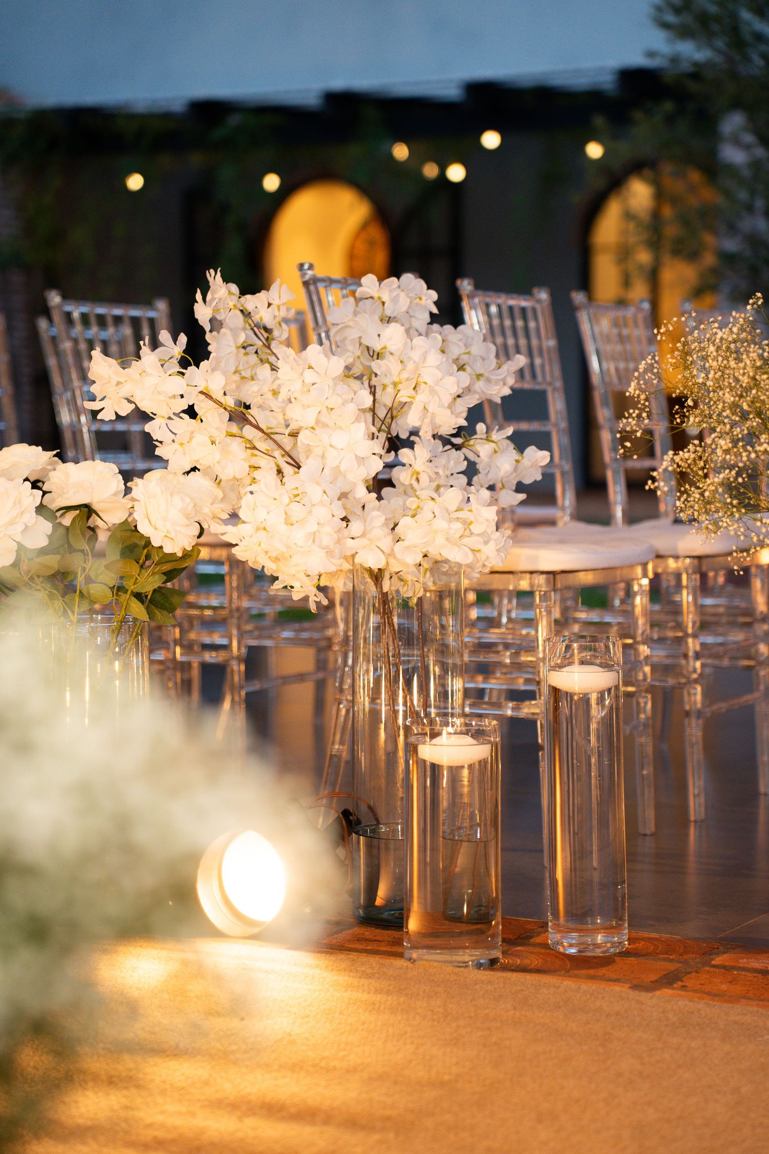 Elegant wedding decor with white floral arrangements, glass candle holders, and glowing lights on a golden aisle at dusk.