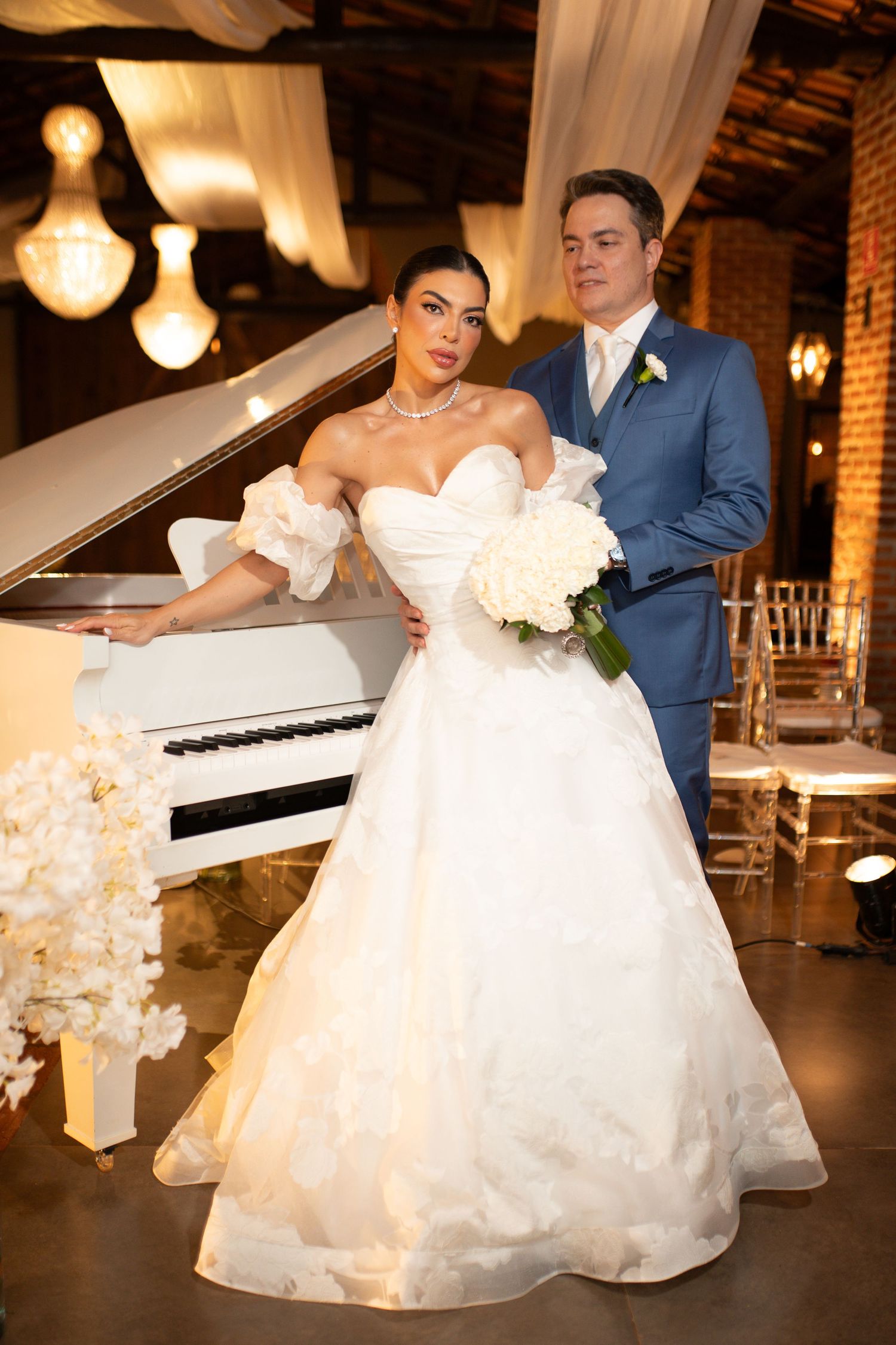 Elegant bride in white ballgown holding bouquet poses with groom in blue suit near white grand piano.