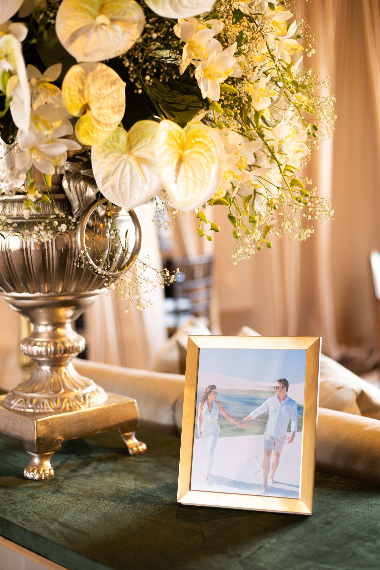 Gold-framed couple photo beside elegant silver urn with white anthurium and lily floral arrangement on green velvet table.