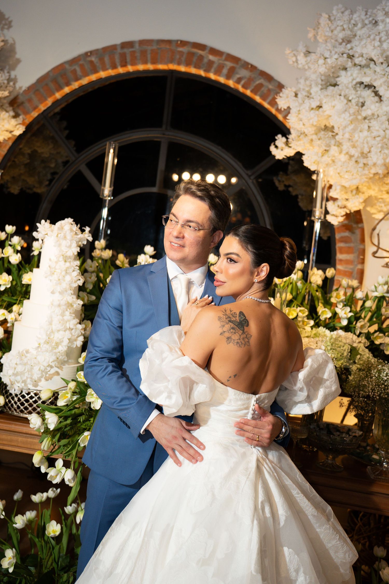 Elegant wedding couple embracing before floral backdrop, bride in off-shoulder gown with tattoo, groom in blue suit.