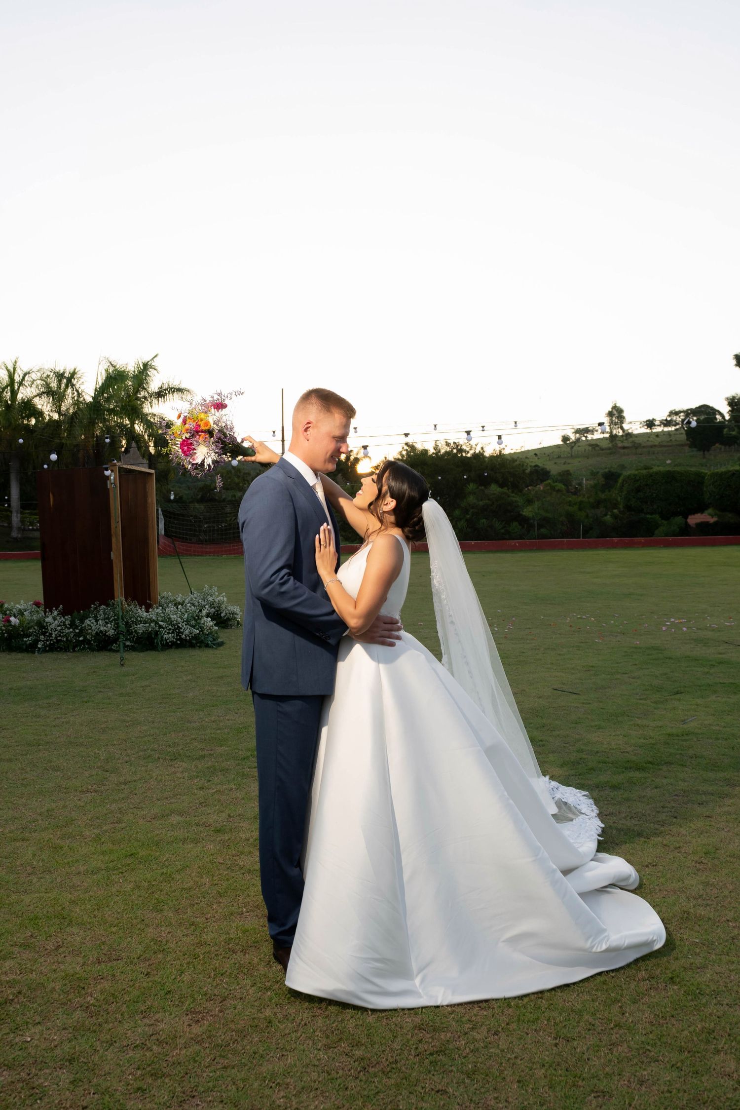Bride in white gown and groom in navy suit share romantic moment on green lawn at outdoor wedding ceremony.