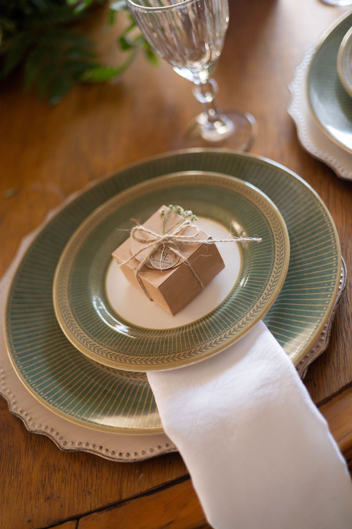 Elegant table setting with sage green plates, kraft paper gift box tied with twine, white napkin, and crystal glass.