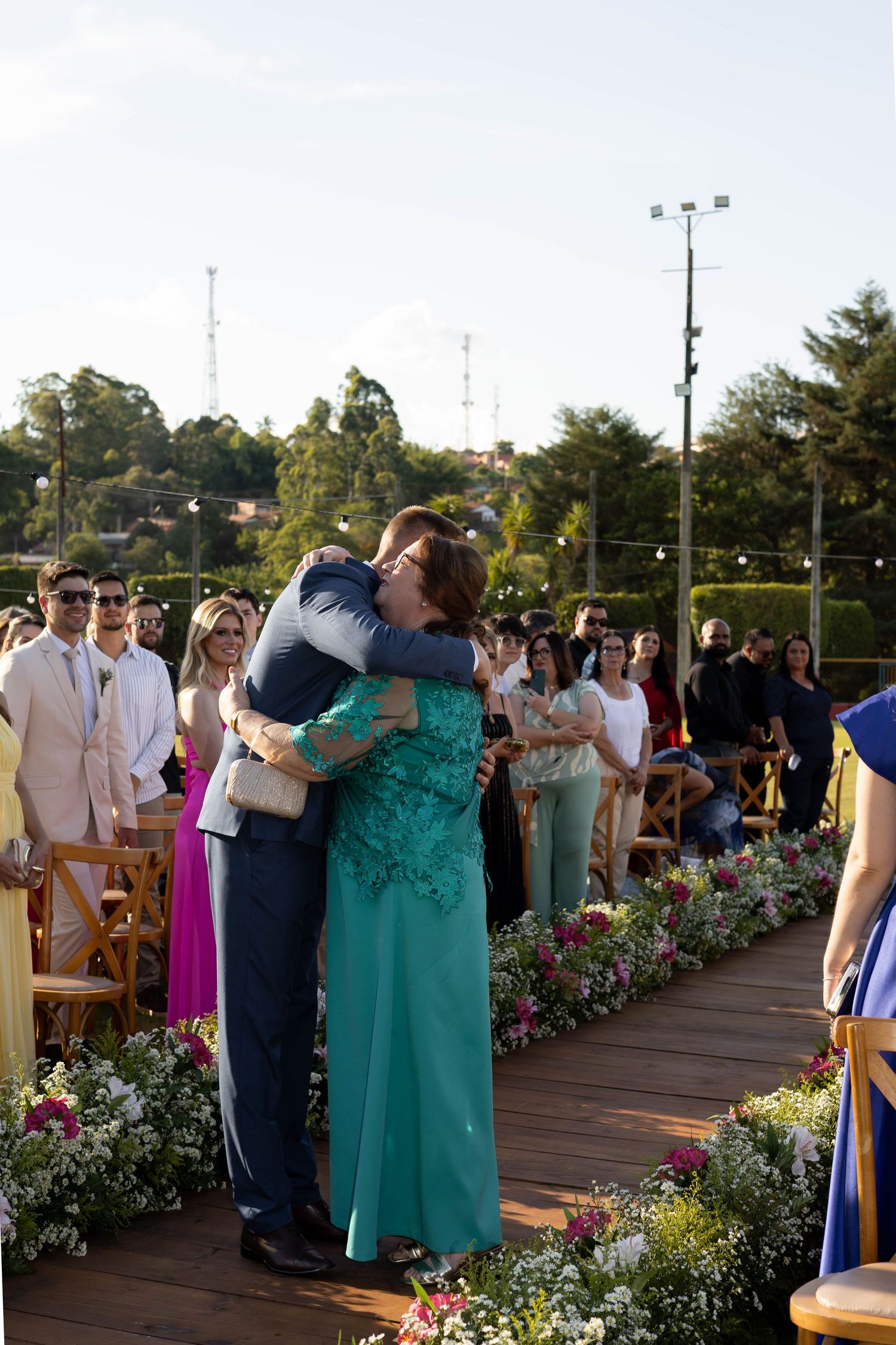 Groom embracing a woman in teal dress at outdoor wedding ceremony with floral aisle and guests watching.