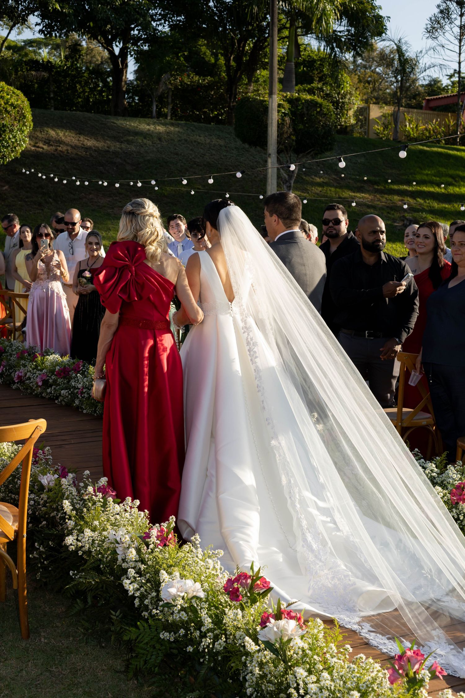 Outdoor wedding ceremony with bride in white gown and veil, groom, and guest in red dress, surrounded by floral arrangements.