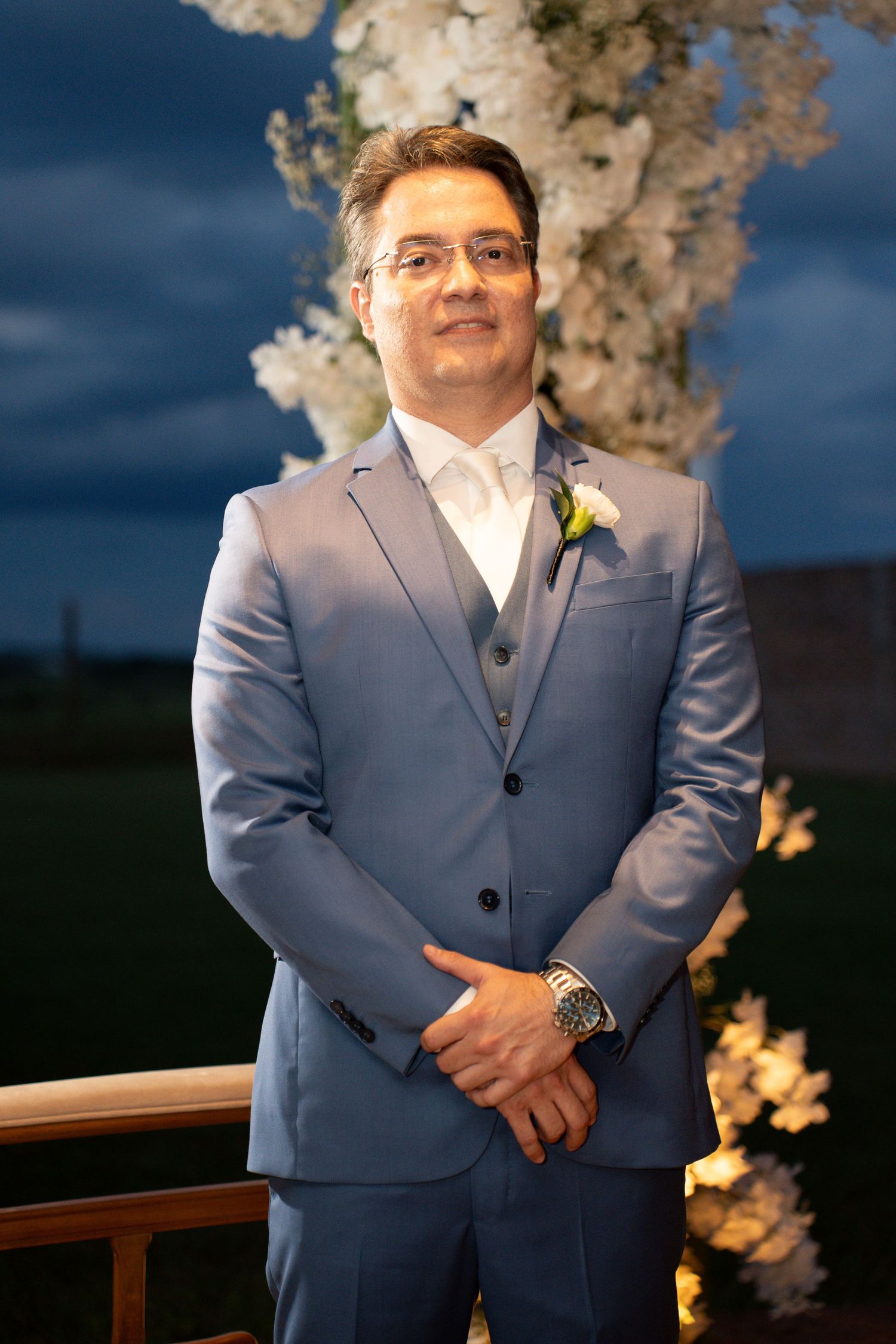 Groom in blue suit with boutonniere stands before white floral arch at outdoor evening wedding ceremony.