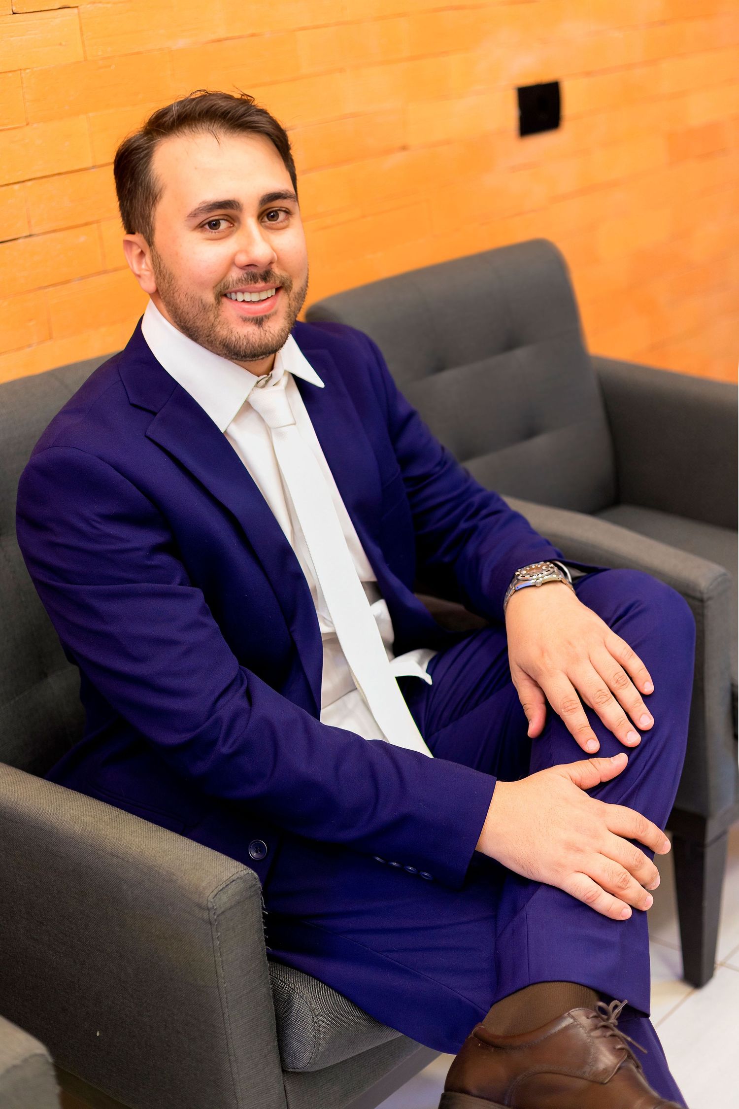 Man in blue suit and tie sitting relaxed in gray armchair, smiling confidently in modern office setting.