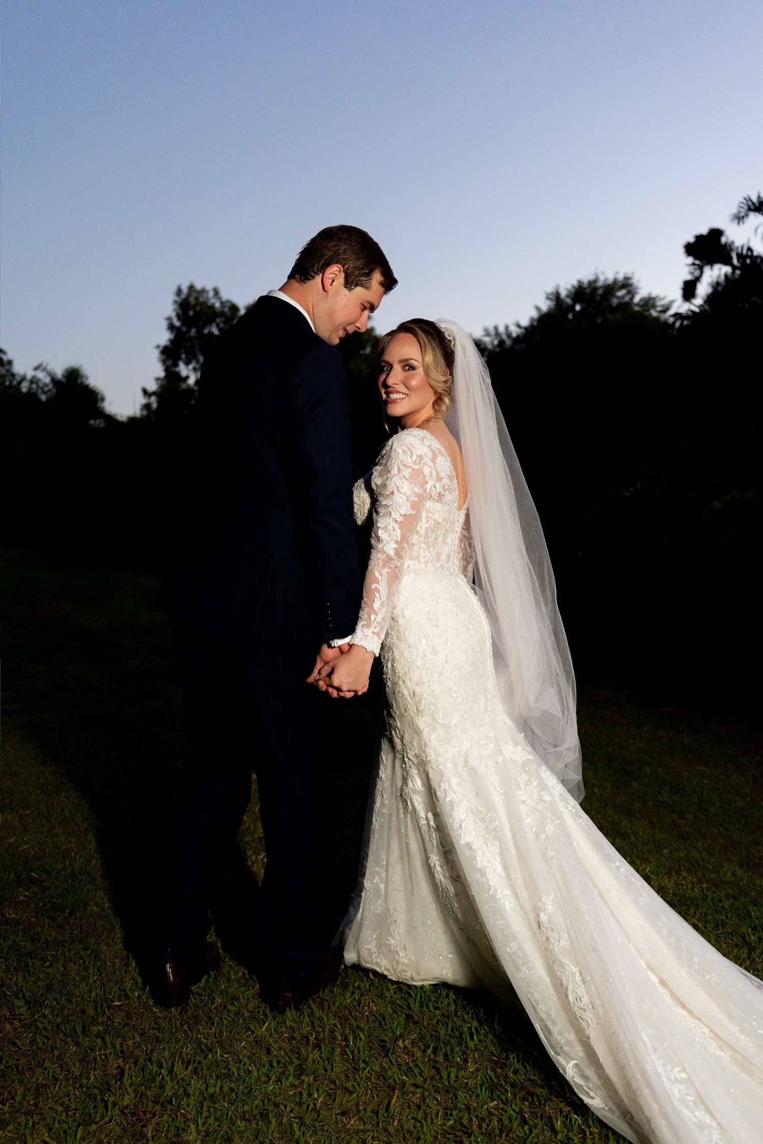 Bride in lace long-sleeve gown and veil holds hands with groom in navy suit at dusk outdoors.