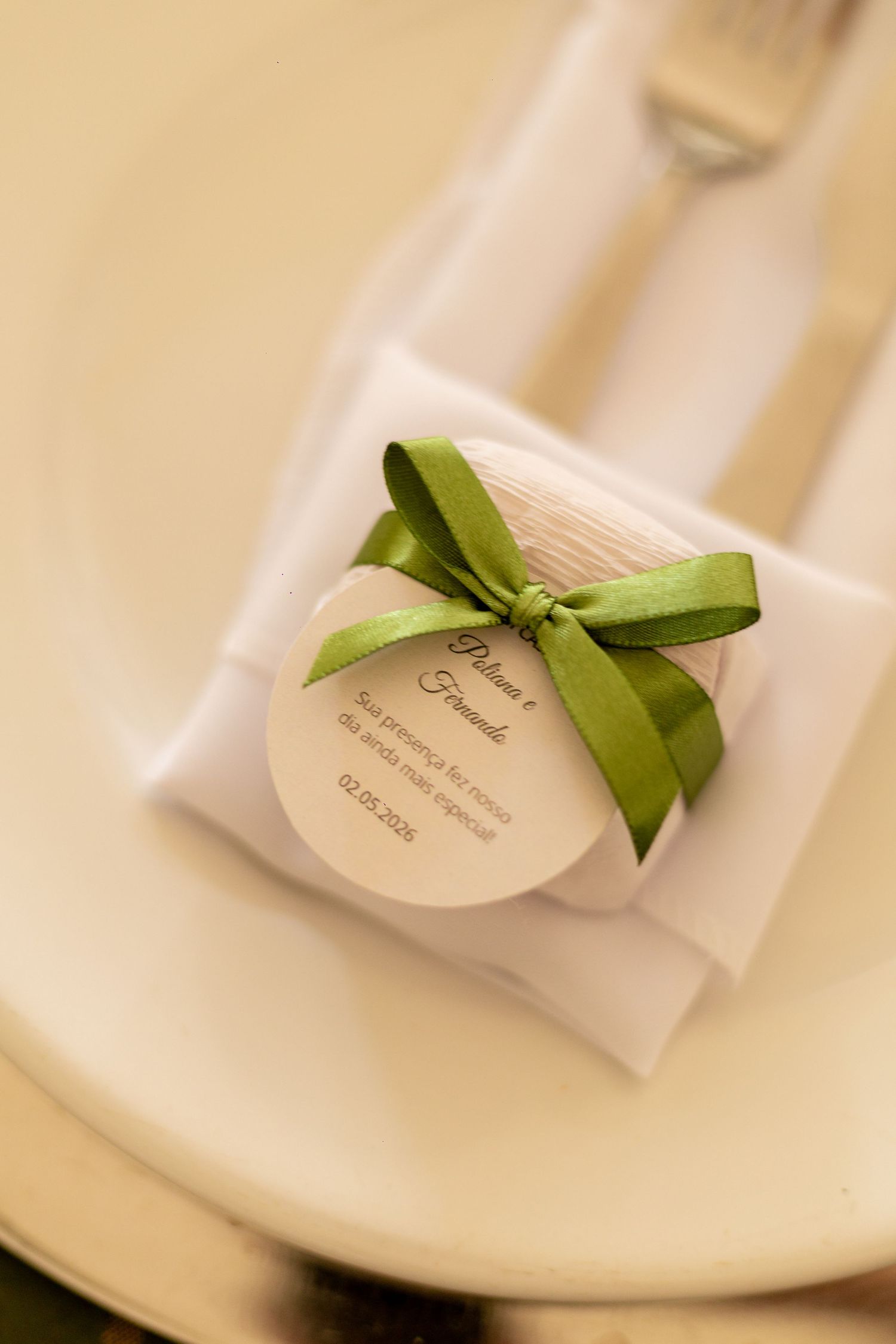 Elegant wedding favor with green ribbon bow on white napkin, personalized round tag on cream plate with silverware.