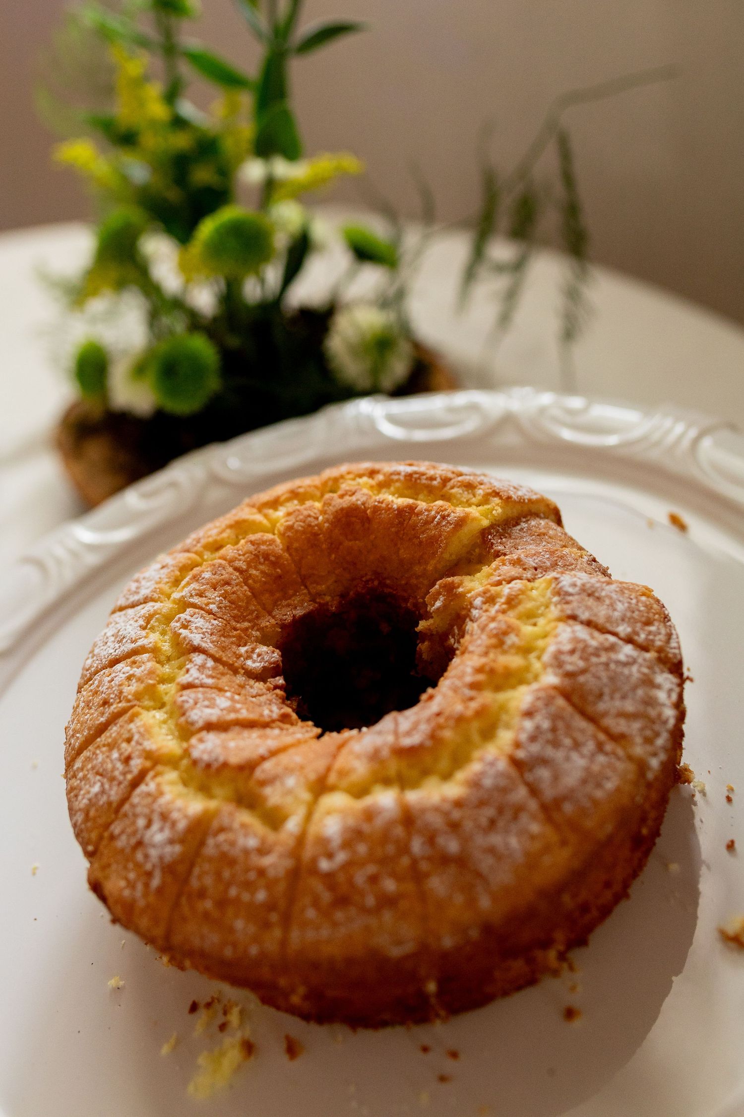 Golden bundt cake dusted with powdered sugar on white plate, with flowers in background.