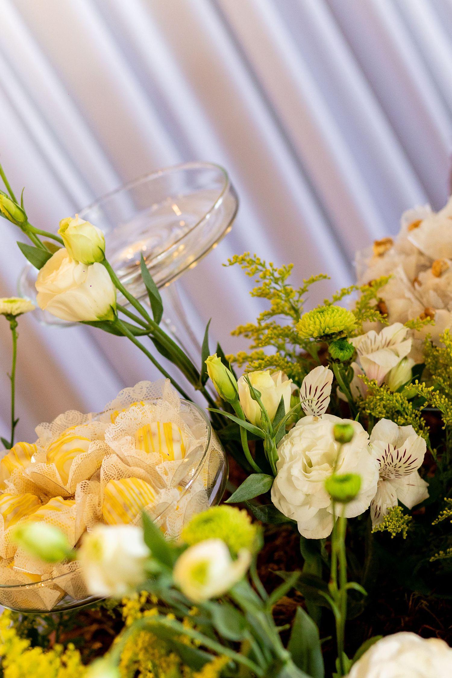 Elegant wedding table decor with white roses, yellow flowers, and glass bowls filled with yellow candies against white draping.