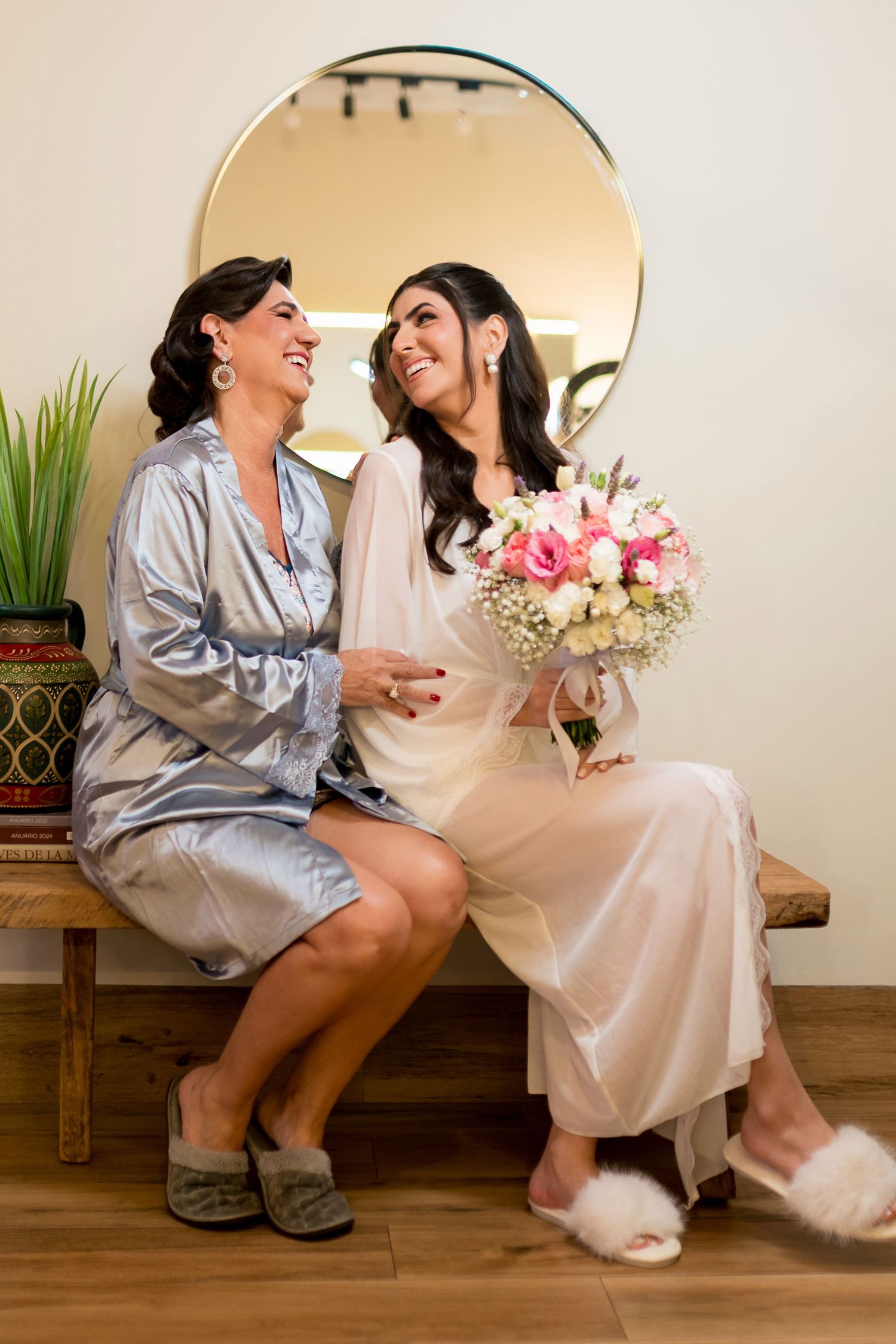 Bride and bridesmaid in satin robes sit on bench laughing, bride holds pink floral bouquet before wedding.