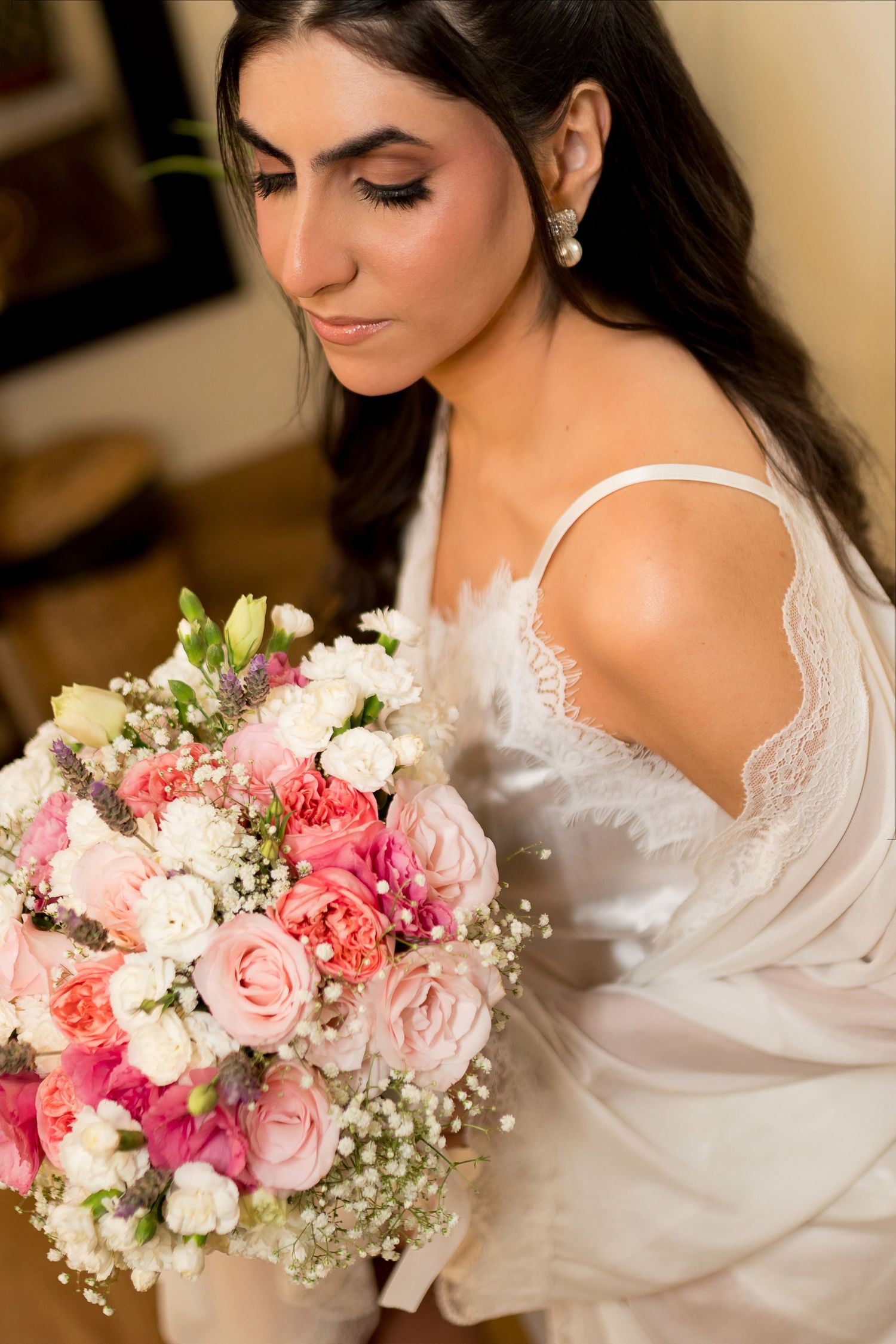 Bride in white lace gown holding a pink and white rose bouquet with baby's breath, looking down elegantly.