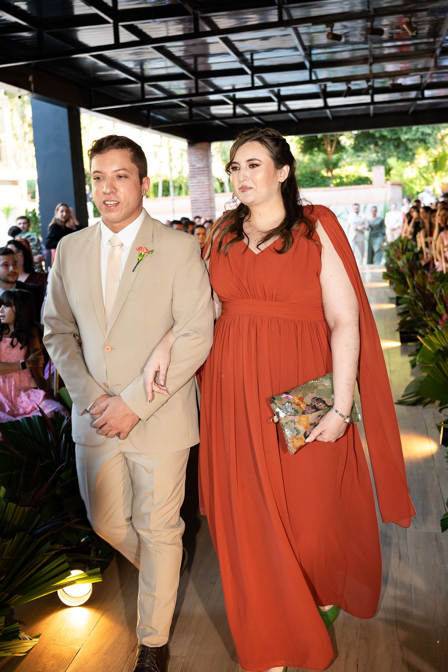 A man in a beige suit and a woman in a rust-red cape gown walk together at a formal wedding event.