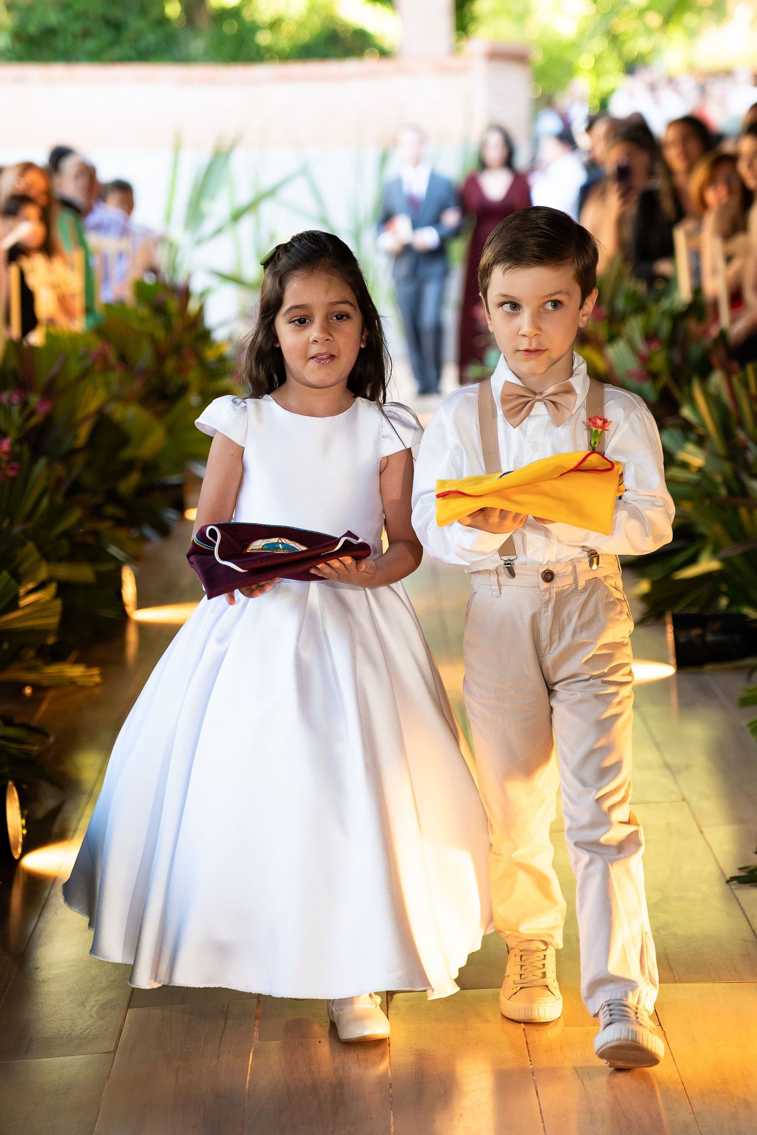 Young flower girl in white dress and ring bearer in suspenders walk down wedding aisle carrying rings and flowers.