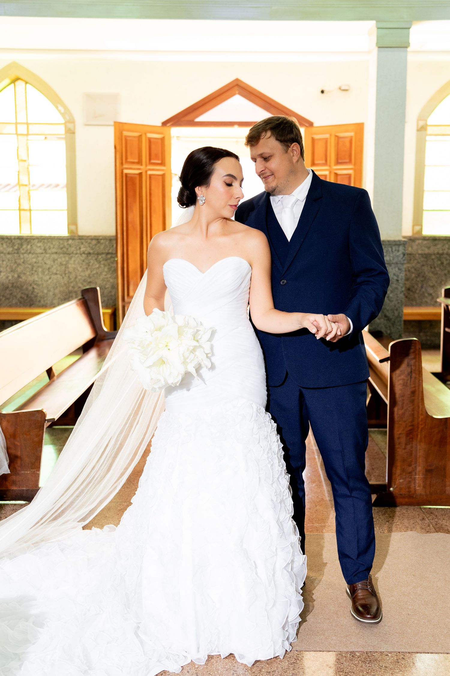 Bride in white strapless gown holding bouquet walks with groom in navy suit down church aisle after wedding ceremony.