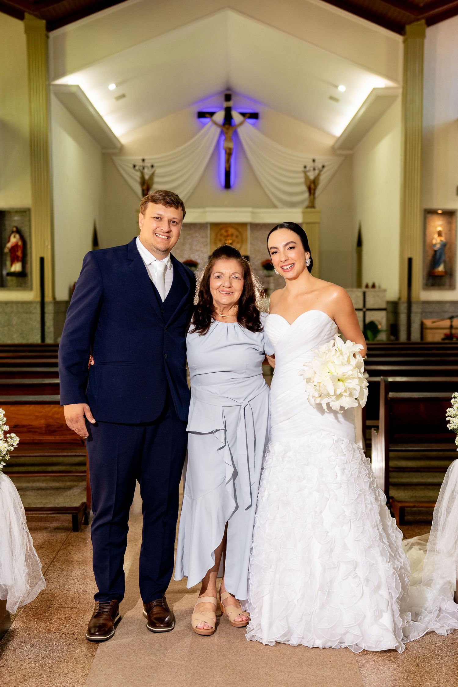 Bride and groom pose with a guest in a decorated Catholic church, bride holding white bouquet.