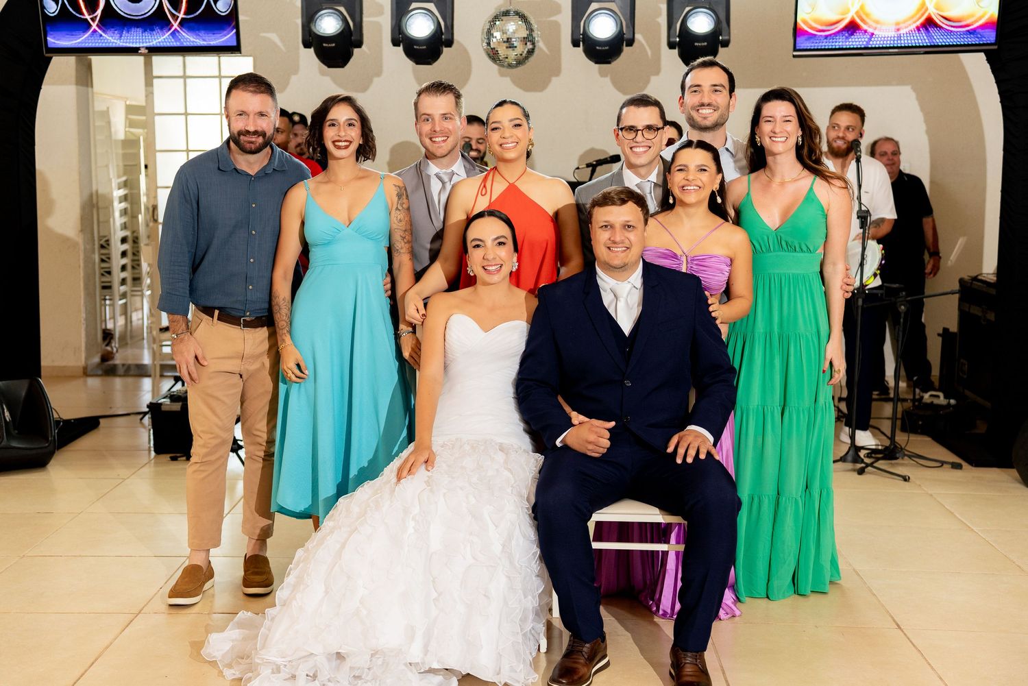 Wedding group photo with bride in white gown and groom in navy suit surrounded by guests in colorful formal attire.