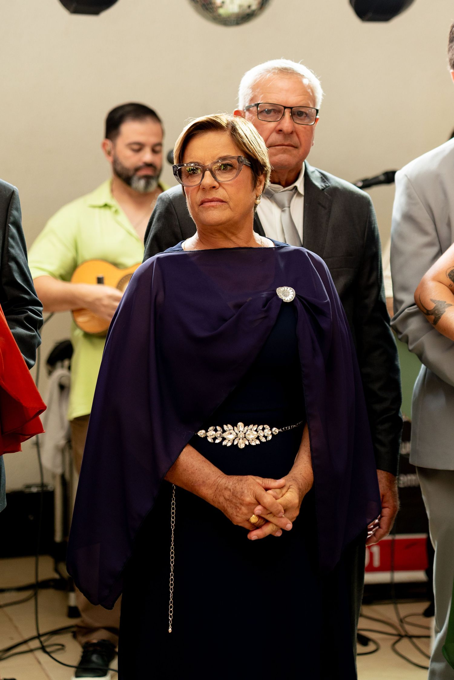 Elegant woman in purple dress with jeweled belt stands at formal event while guitarist performs in background.