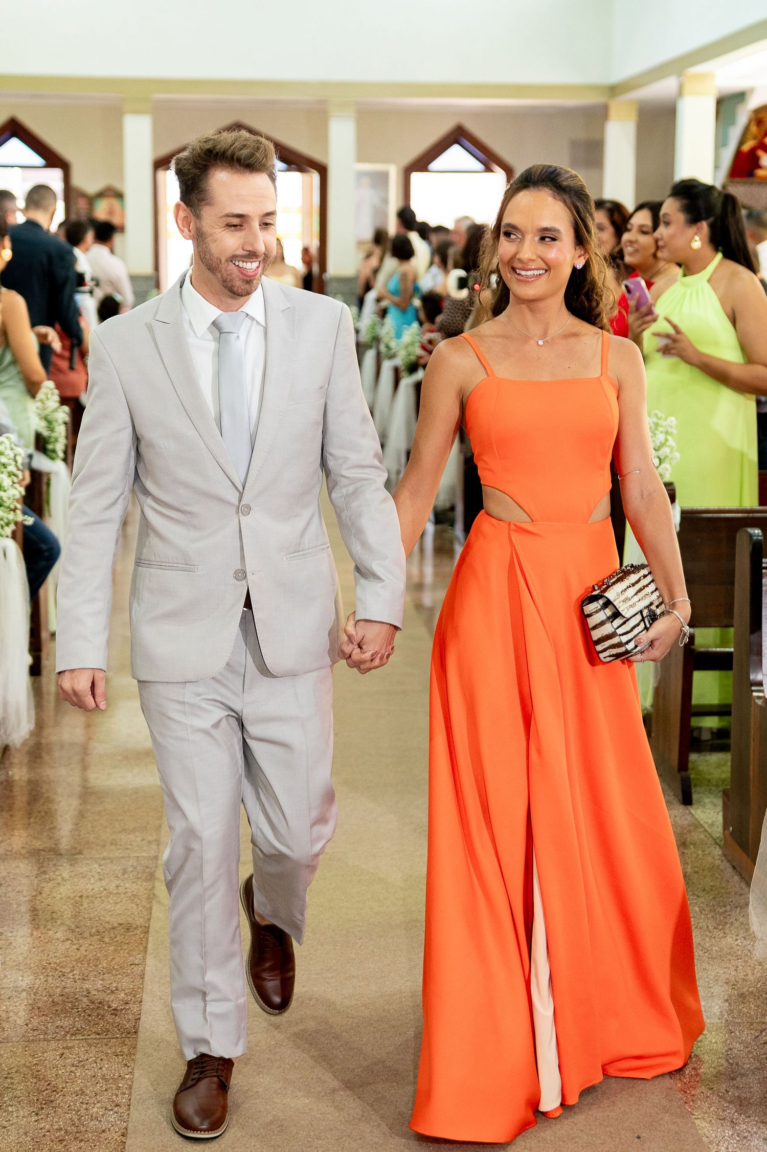 Smiling couple walks down church aisle, man in gray suit, woman in orange gown holding clutch at wedding.