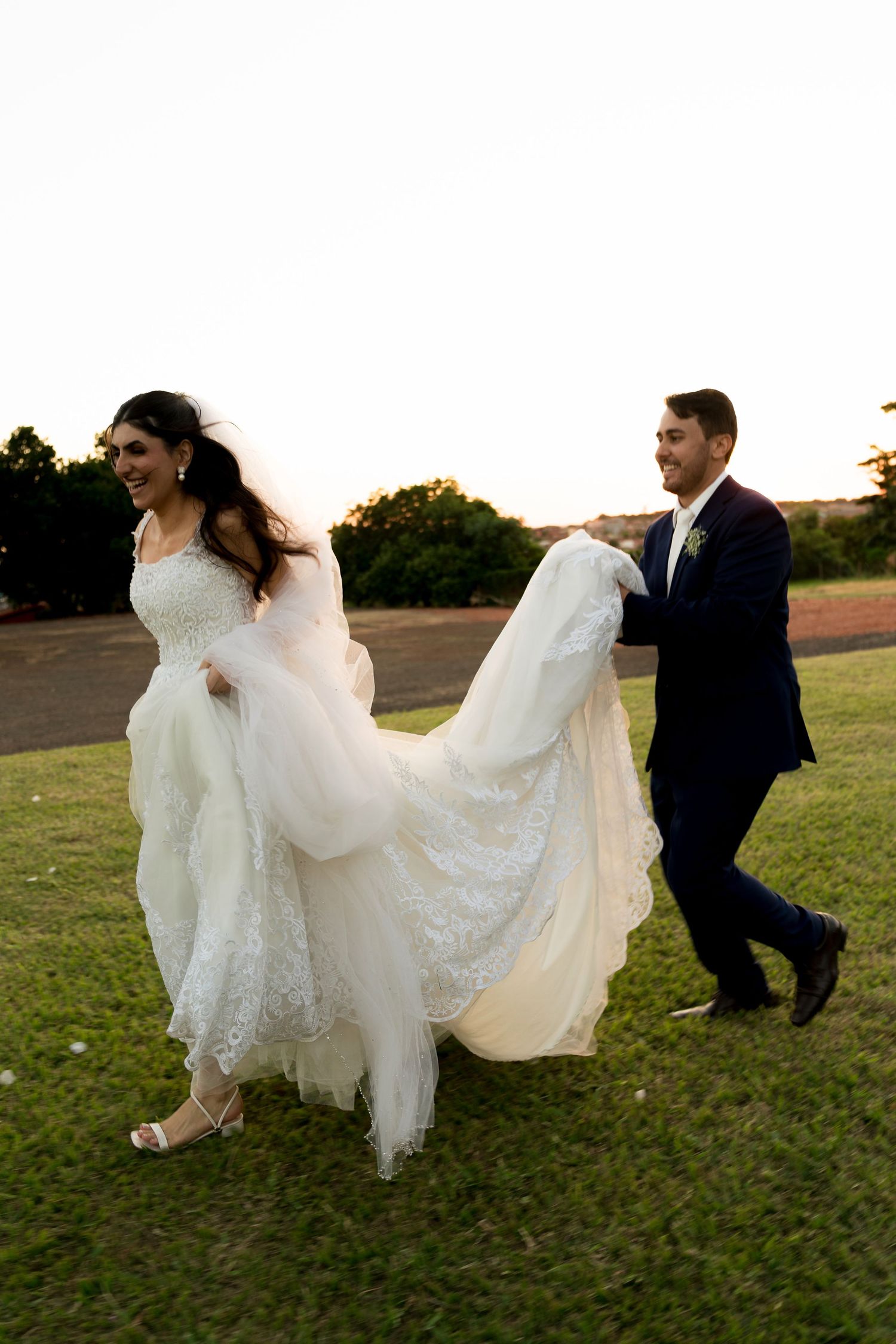 A joyful bride in a white lace gown runs across green grass as her groom in a navy suit carries her train at sunset.